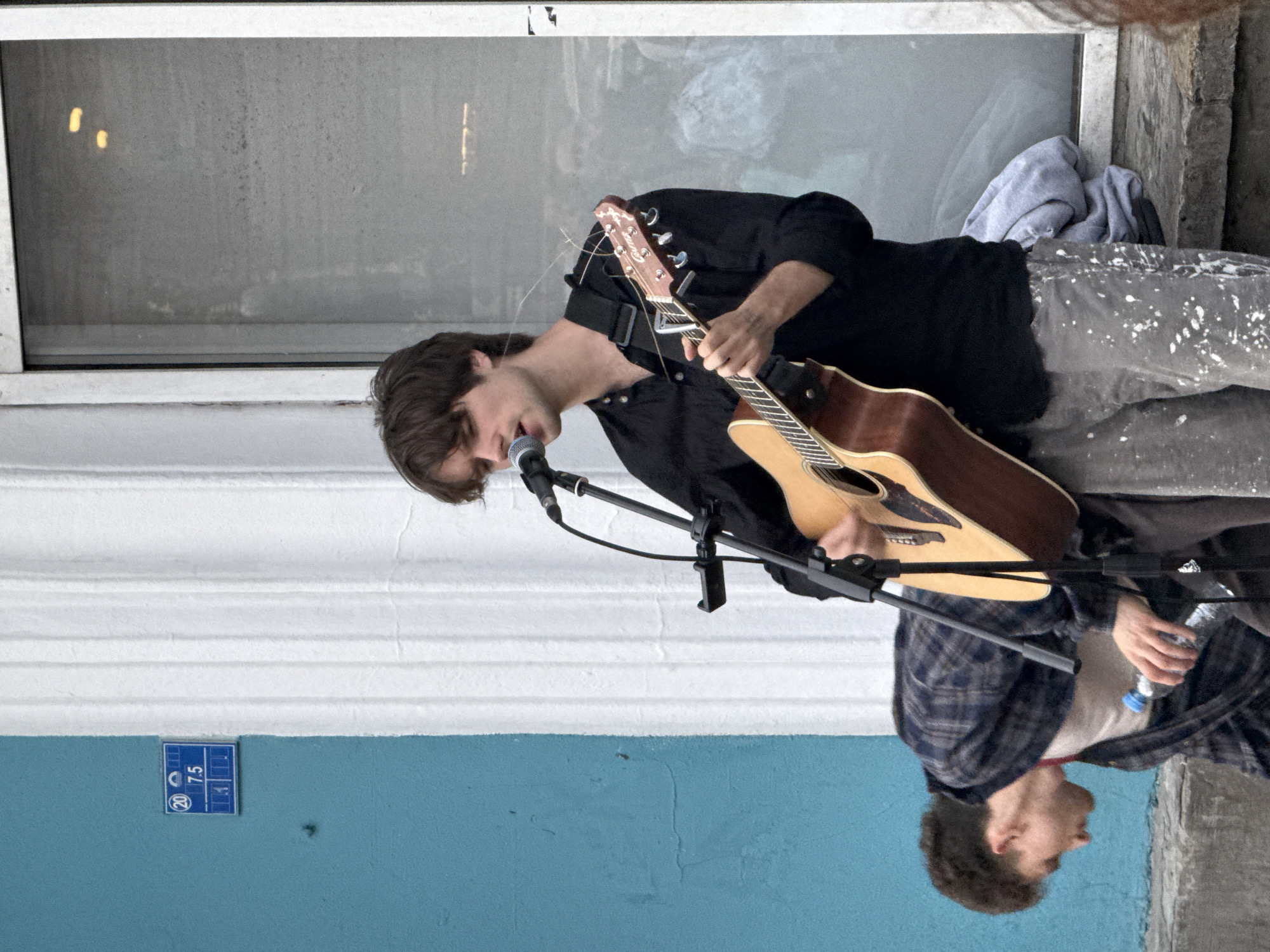In Saint Petersburg, Russia, a young man with dark hair is captured mid-performance, singing into a microphone while playing an acoustic guitar. He is wearing a black button-down shirt and grey pants speckled with white paint. To his left, another young man in a plaid shirt is bending down, holding a water bottle. They are positioned in front of a white columned structure and a blue wall. A small blue sign with white text and numbers, possibly indicating a house or street number, is affixed to the blue wall. The background appears to be a street scene with reflections in a window.