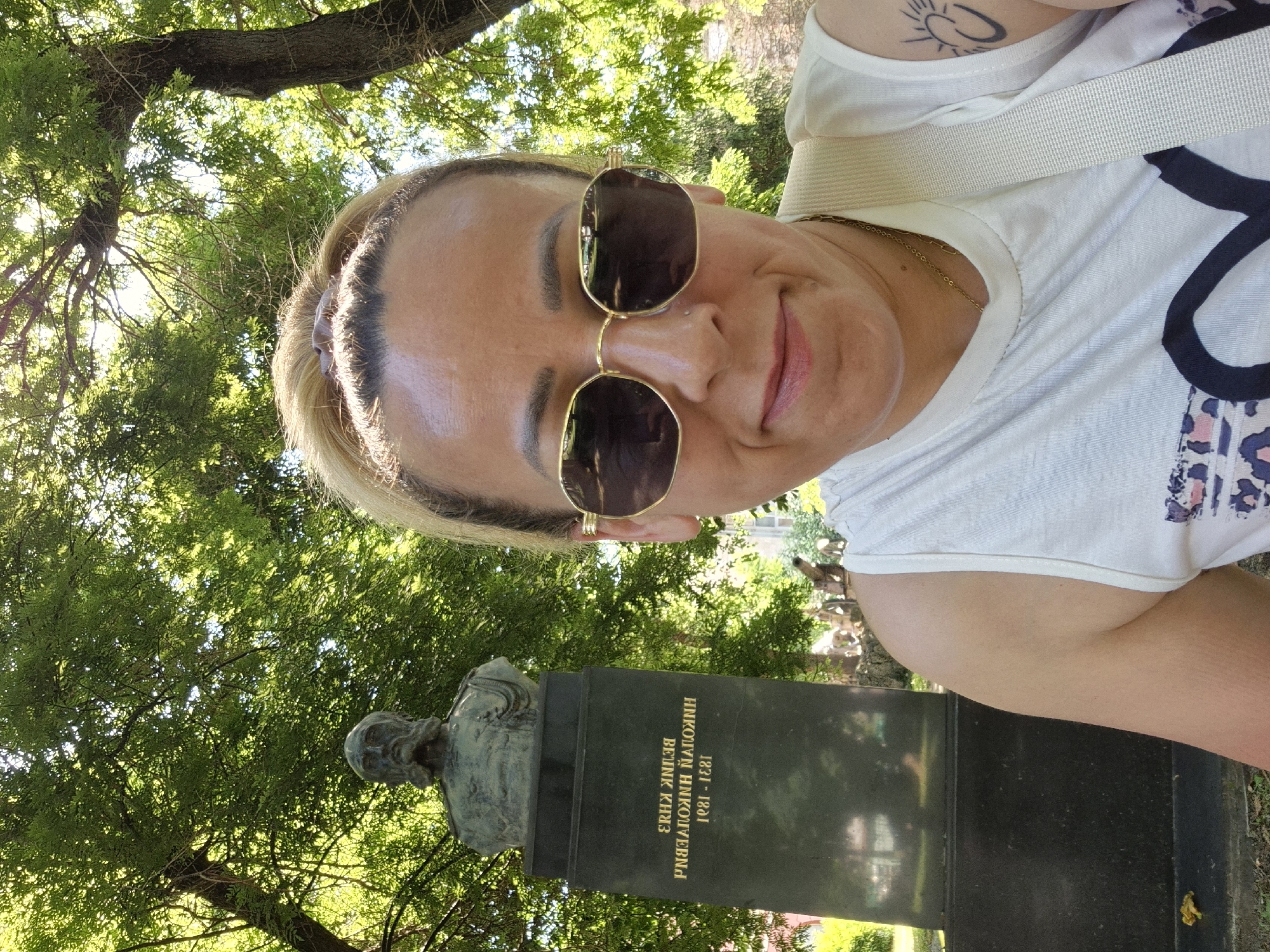 A woman with blonde hair pulled back and wearing sunglasses, a white tank top, and a gold necklace poses for a selfie in front of a monument in a park. The monument is a dark stone pedestal with a bust of a man on top, and text inscribed on the front. The woman has a tattoo on her right arm. The background is filled with lush green trees and dappled sunlight. In the distance, other people and some historical artifacts like cannons are visible. The image is taken outdoors during the daytime.