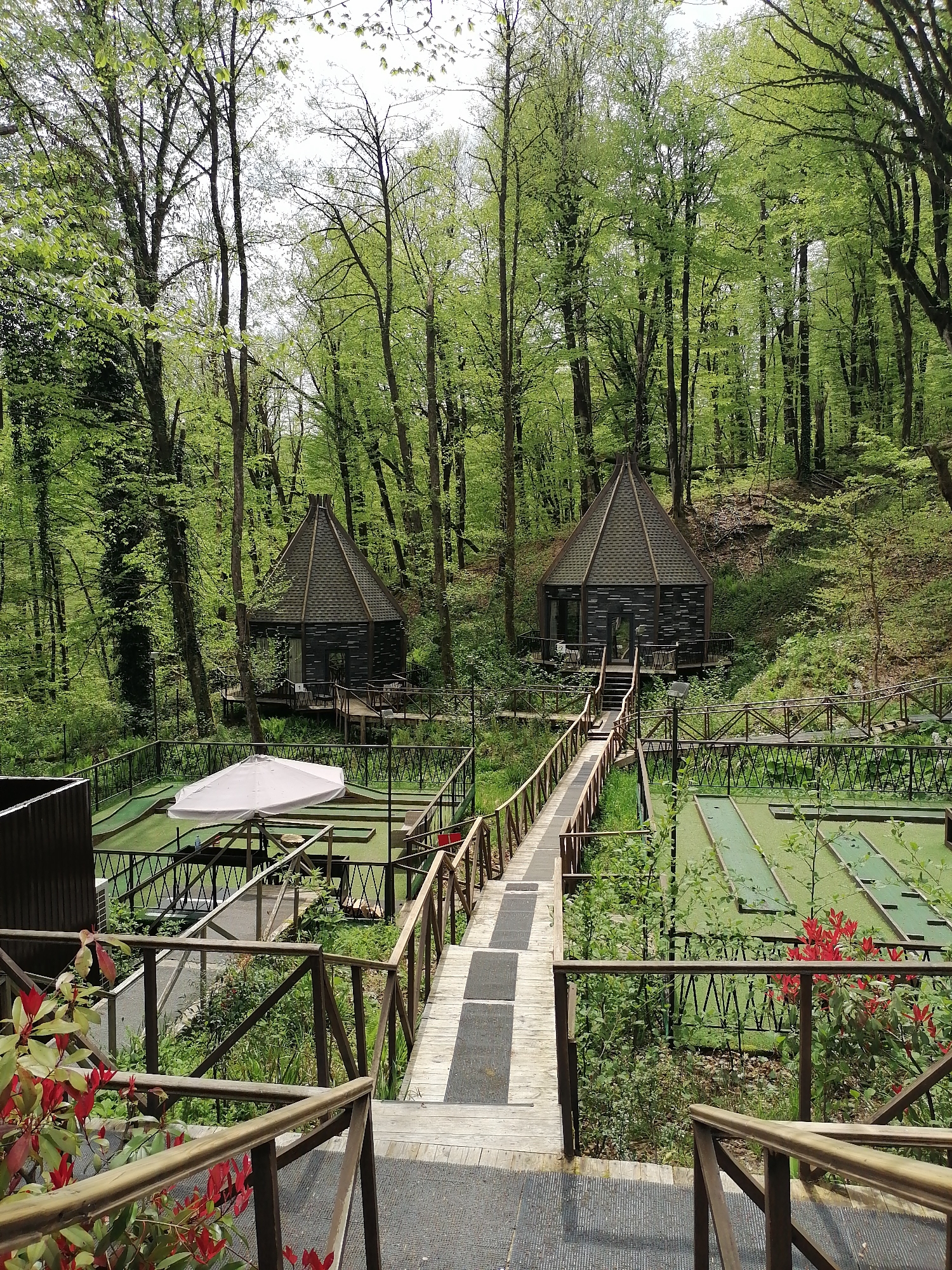 The image depicts a recreational area in Medoveevka, Russia, nestled within a forest. Two small, dark wooden cabins with peaked roofs are visible in the background, situated on platforms and connected by a wooden bridge. In the foreground, a mini-golf course is enclosed by fencing. An umbrella shades a section of the course. The view is from an elevated position, looking down a set of steps with wooden railings toward the scene. Red flowering plants add color to the foreground. The surrounding environment is lush with green trees and vegetation.