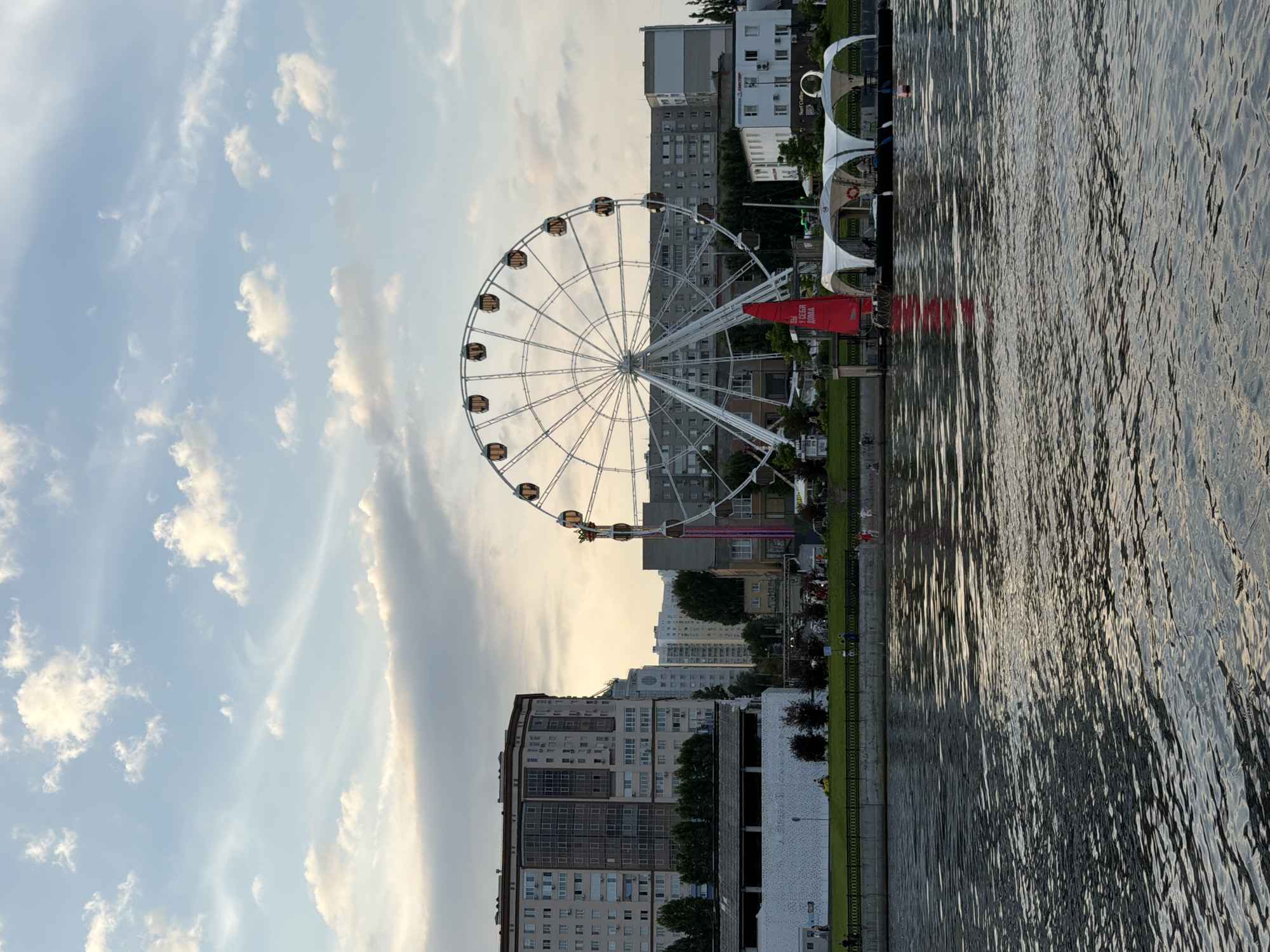 The image captures a view of the Saratov Embankment along the Volga River in Saratov, Russia, at dusk. A large Ferris wheel is prominently featured in the center of the frame, with several gondolas visible. In the foreground, the rippling surface of the Volga River reflects the soft light of the setting sun. The middle ground shows a section of the embankment with people walking and relaxing. Several buildings, including apartment blocks and a white structure, are visible behind the embankment. A red sail, possibly attached to a boat or part of a decorative structure, is also present. The sky is partly cloudy with a mix of blue and diffused light. The overall scene depicts a peaceful urban waterfront setting.