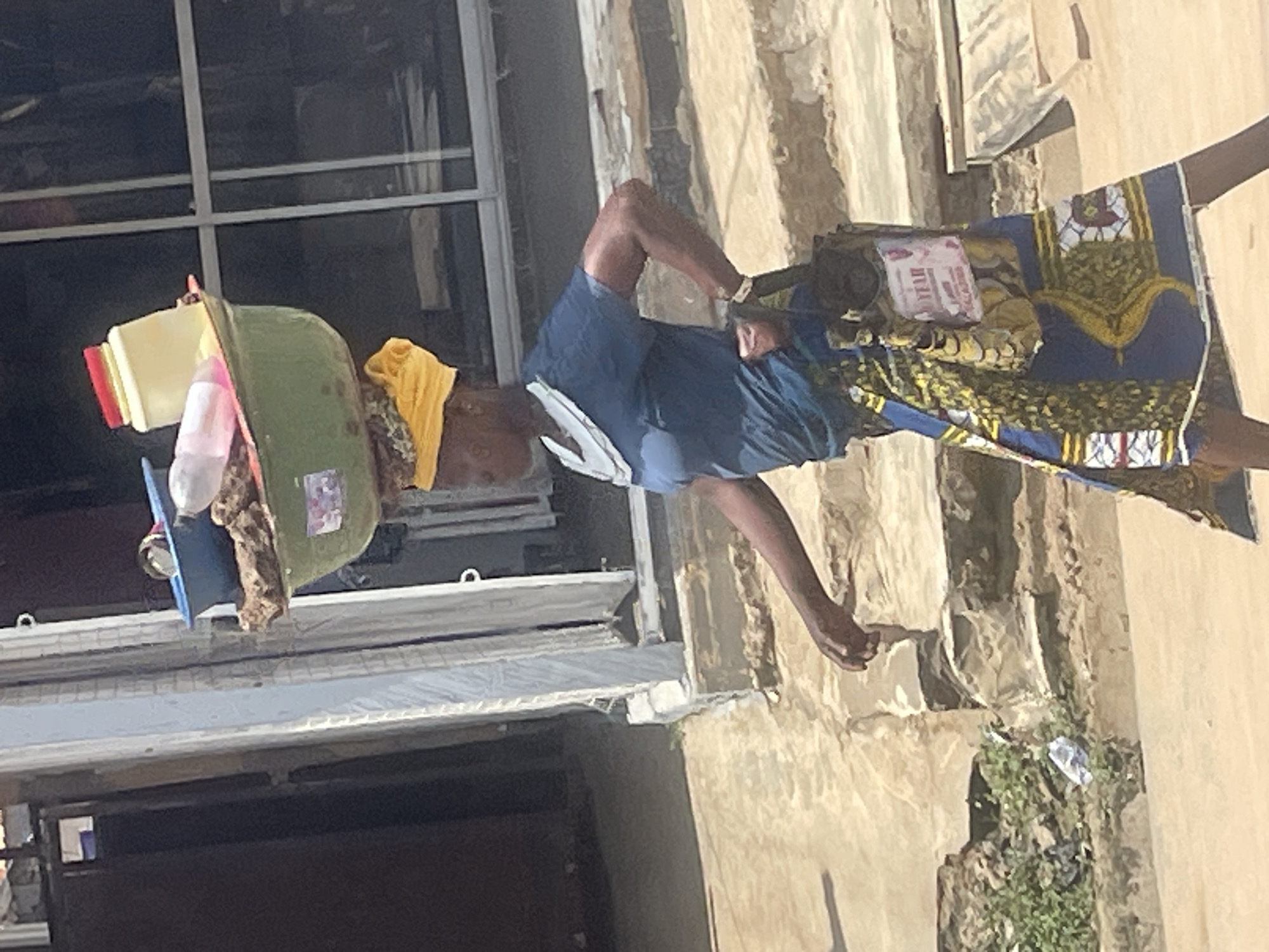 A woman is walking outdoors, balancing a large green basin filled with items on her head. She is wearing a blue t-shirt, a yellow headscarf, and a colorful patterned skirt. A bag is hanging from her arm. The background shows a building with windows and a stepped sidewalk. The scene appears to be in an urban environment, possibly the city of Abuja, Nigeria, given the presence of vendors often seen in such markets. The lighting suggests it is daytime.