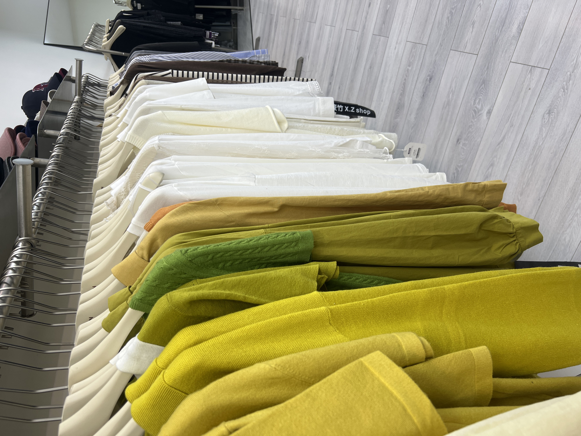 The image shows a clothing store in Osh City, Kyrgyzstan, with racks of women's clothing. The clothing is mostly in muted earth tones and pastel colors, with a few brighter pieces. The store appears to be well-organized and clean. The floor is a light gray wood grain laminate. There are no people visible in the image.