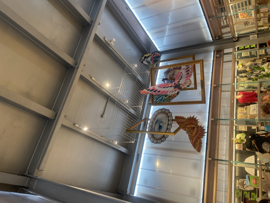 This image depicts the interior of a retail space, specifically identified as Liberty London, located on Regent Street, City of Westminster, London, United Kingdom.
The scene focuses on the upper section of the store, featuring an industrial-style ceiling with exposed grey metal beams and a light grey finish. Recessed spotlights and track lighting are visible, along with a white smoke detector. Suspended from a metal grid structure attached to the ceiling are multiple decorative art installations.
Prominently featured is a large, oval-shaped eye with a visible tear, encircled by faux pearls, connected to a prominent orange and red butterfly wing, all contained within a geometric gold-colored frame structure. To its immediate right, a pair of vibrant pink, green, and black butterfly wings are superimposed over a partial human face, framed by a rectangular gold frame. A smaller blue, black, and yellow butterfly wing hangs further to the right. These installations are suspended by thin wires.
Below the ceiling and installations, the upper wall sections consist of translucent, ribbed panels, backlit with a cool-white light. Beneath these panels, a retail area is partially visible, displaying wooden shelving units. Mannequins are present, one wearing a black garment with an eye motif, and another in a red blouse. Various retail merchandise, including books, vases, and other decorative items, are arranged on shelves and displays. In the lower right foreground, the top of a person's head with brown, curly hair is partially visible and out of focus, indicating human presence within the store.
