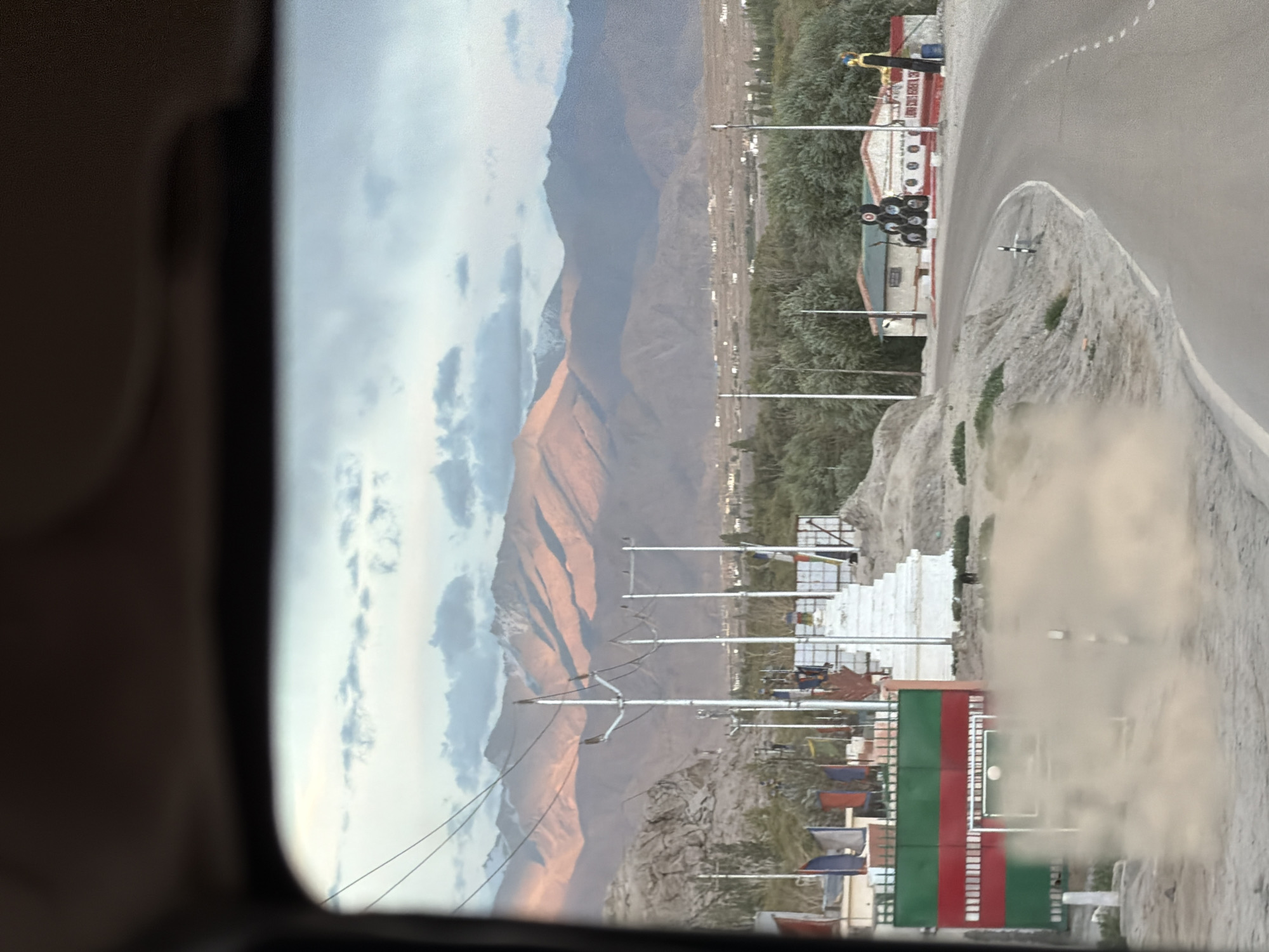 The image depicts a scenic view of a mountainous landscape with a road curving through it. The foreground features a dusty road and a vibrant green and red striped fence. Behind the fence, prayer flags flutter in the wind, and a white stupa stands prominently. In the middle ground, a building with colorful decorations and stacked tires is visible. The background is dominated by majestic mountains, bathed in the warm light of what appears to be sunrise or sunset. The sky is partly cloudy, with a mix of blue and white. The scene is suggestive of the region around Spituk, India, known for its monasteries and dramatic mountain scenery. There are no people clearly visible in the image, though a small black object that could be an animal is present near the fence. The overall impression is one of natural beauty and cultural significance.