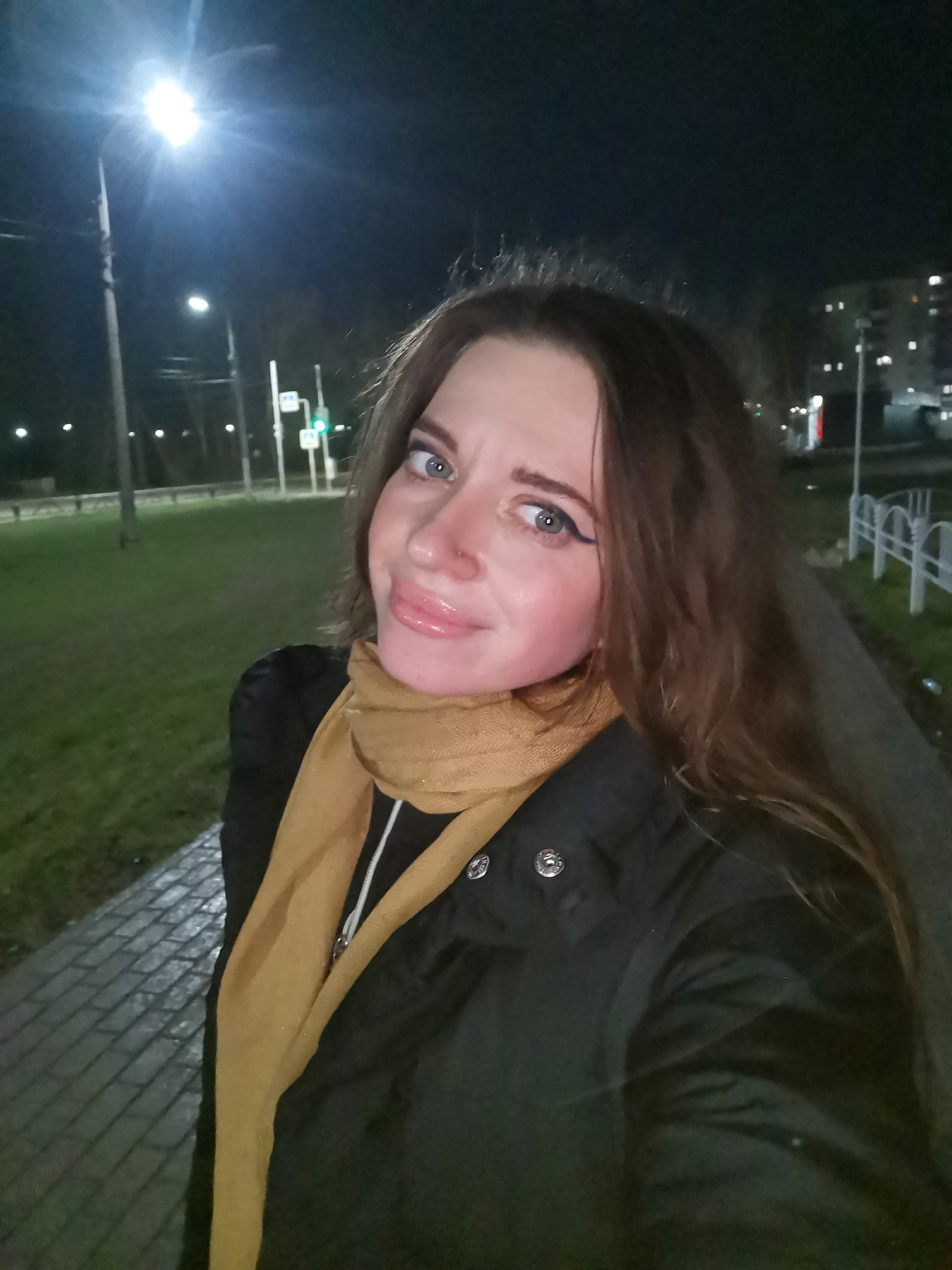 A woman poses for a selfie outdoors at night. She has long brown hair, light eyes with dark eyeliner, and is wearing a black jacket and a mustard-colored scarf. The background features streetlights and a grassy area with a sidewalk. A building with illuminated windows is visible in the distance.