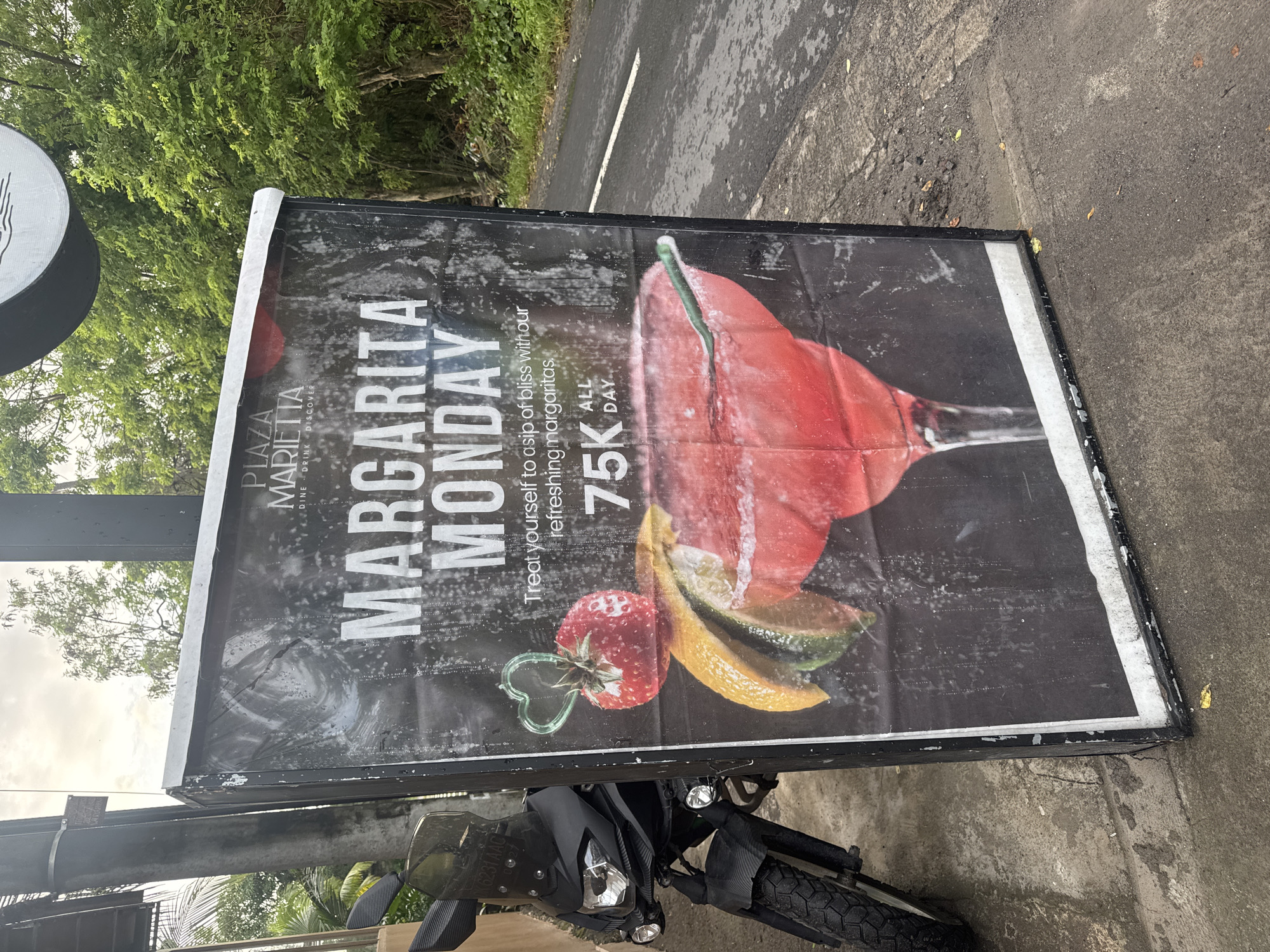 The image shows a promotional sign for "Margarita Monday" at "Plaza Marietta," located in Nusa Penida, Indonesia. The sign advertises margaritas for 75K all day. A motorcycle is parked in front of the sign, partially obscuring the bottom. The sign's background is black, with a large image of a red margarita, a strawberry with a heart-shaped stem, and citrus fruit slices. The sign is placed near a road and some foliage is visible in the background.