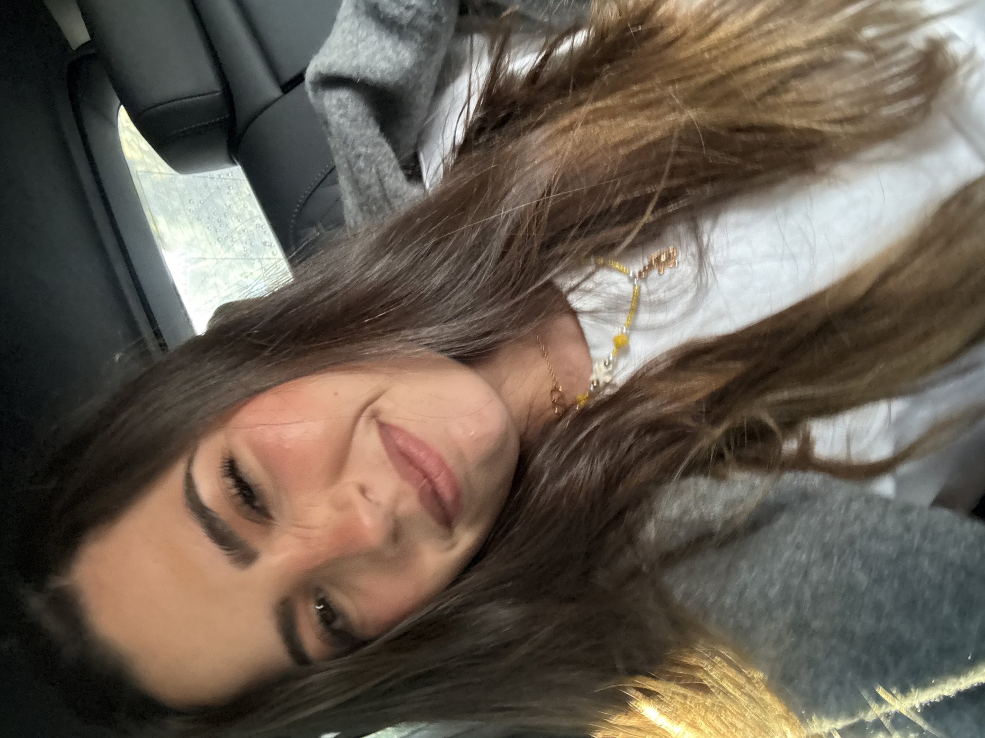 A selfie portrait of a woman with long brown hair, fair skin, and dark eyebrows. She is smiling slightly. She is wearing a white shirt, a gray sweater, and a gold-colored necklace. She appears to be in the back seat of a car, with black leather seats visible. A window can be seen, with what appears to be a rainy day outside. There is no indication of the specific location in the city of Puebla, Mexico.
