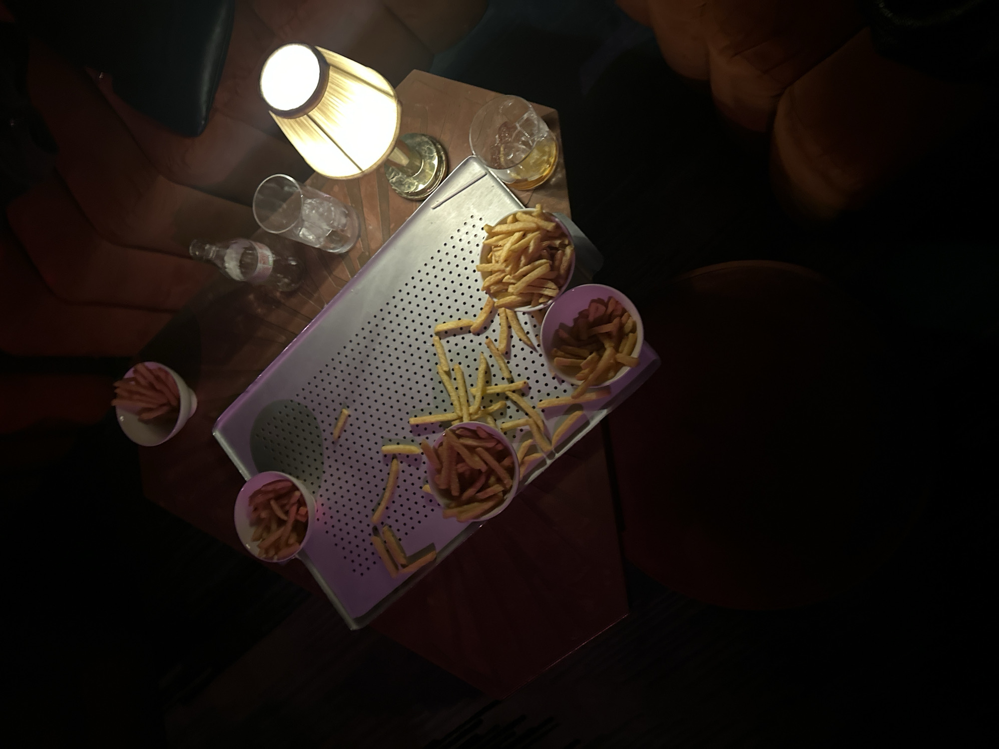 A high-angle shot reveals a dimly lit bar or lounge in what appears to be London, United Kingdom. In the left side of the frame, portions of chairs and sofas are visible, upholstered in dark and orange materials. A small table made of what appears to be wood is illuminated by a lamp. A perforated metal tray holds five small bowls of french fries and several individual fries are scattered on the tray. There are also two empty glasses, a bottle of water and a glass of what appears to be whiskey.