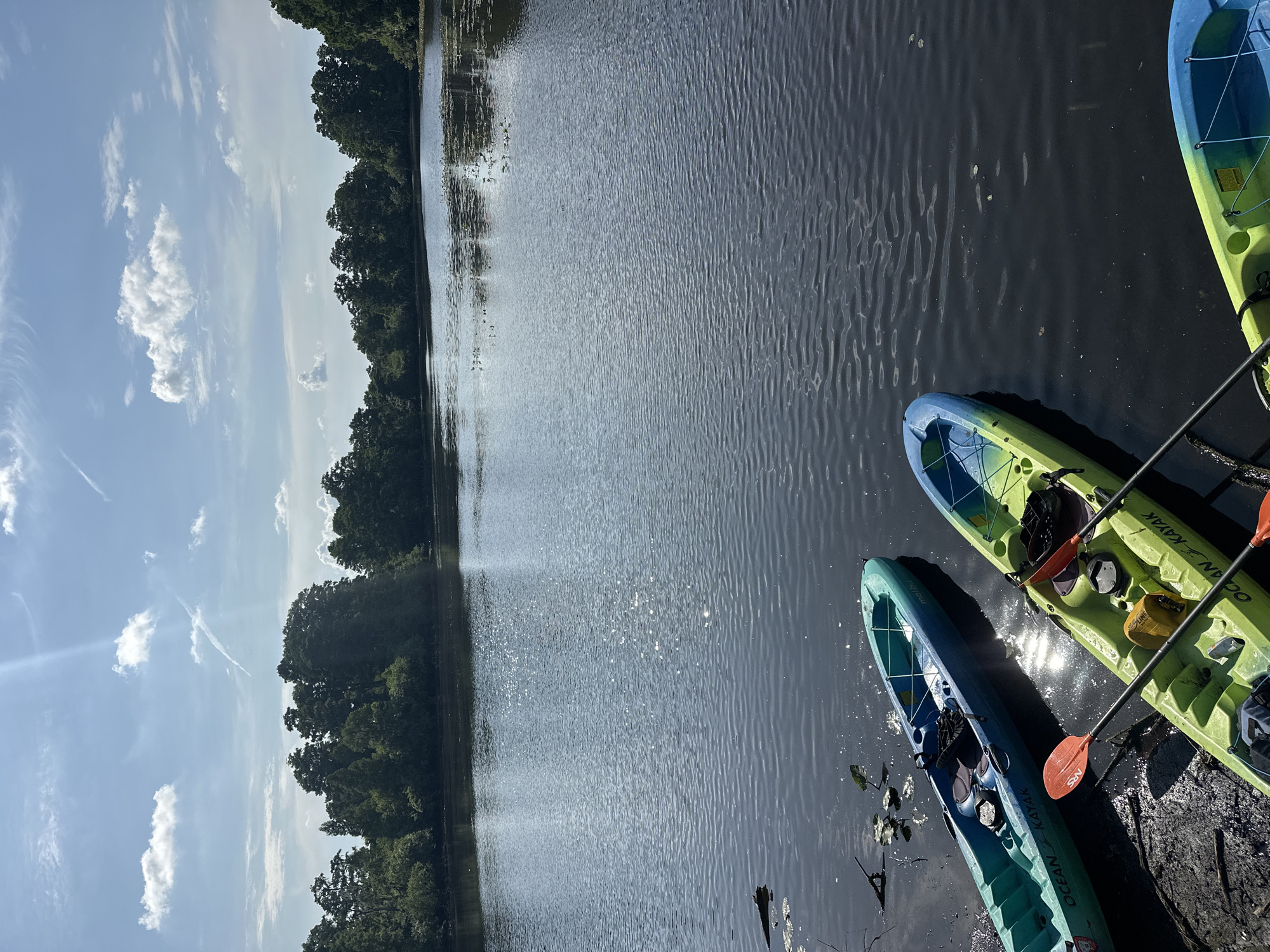 The image shows a body of water, possibly a lake or wide river, reflecting sunlight. Several kayaks are docked on the right side of the frame. The sky is partly cloudy with scattered clouds. Trees line the edge of the water on the left side of the image. There are no people visible. It is not possible to identify the precise location in Tampa with the data provided.