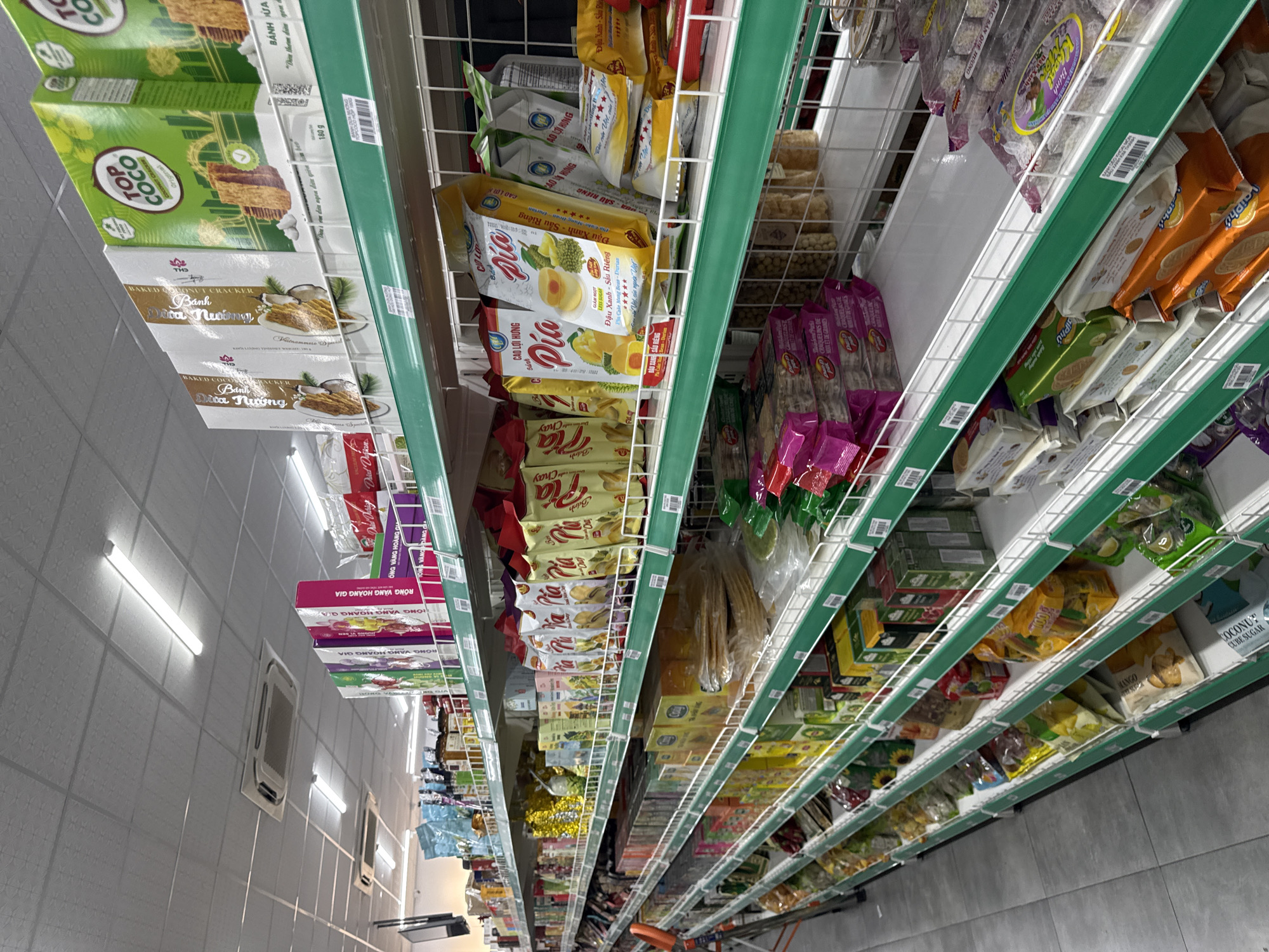 The image shows a retail shelf aisle in a store, likely in Đà Nẵng, Vietnam, based on the Vietnamese language on the product packaging. The shelves are stocked with various food products including coconut crackers, "Pia" pastries, and other snacks. The shelves have green trim. The aisle is illuminated by fluorescent lighting on the ceiling. The perspective suggests the photo was taken from a low angle, possibly from the floor or a shopping cart.