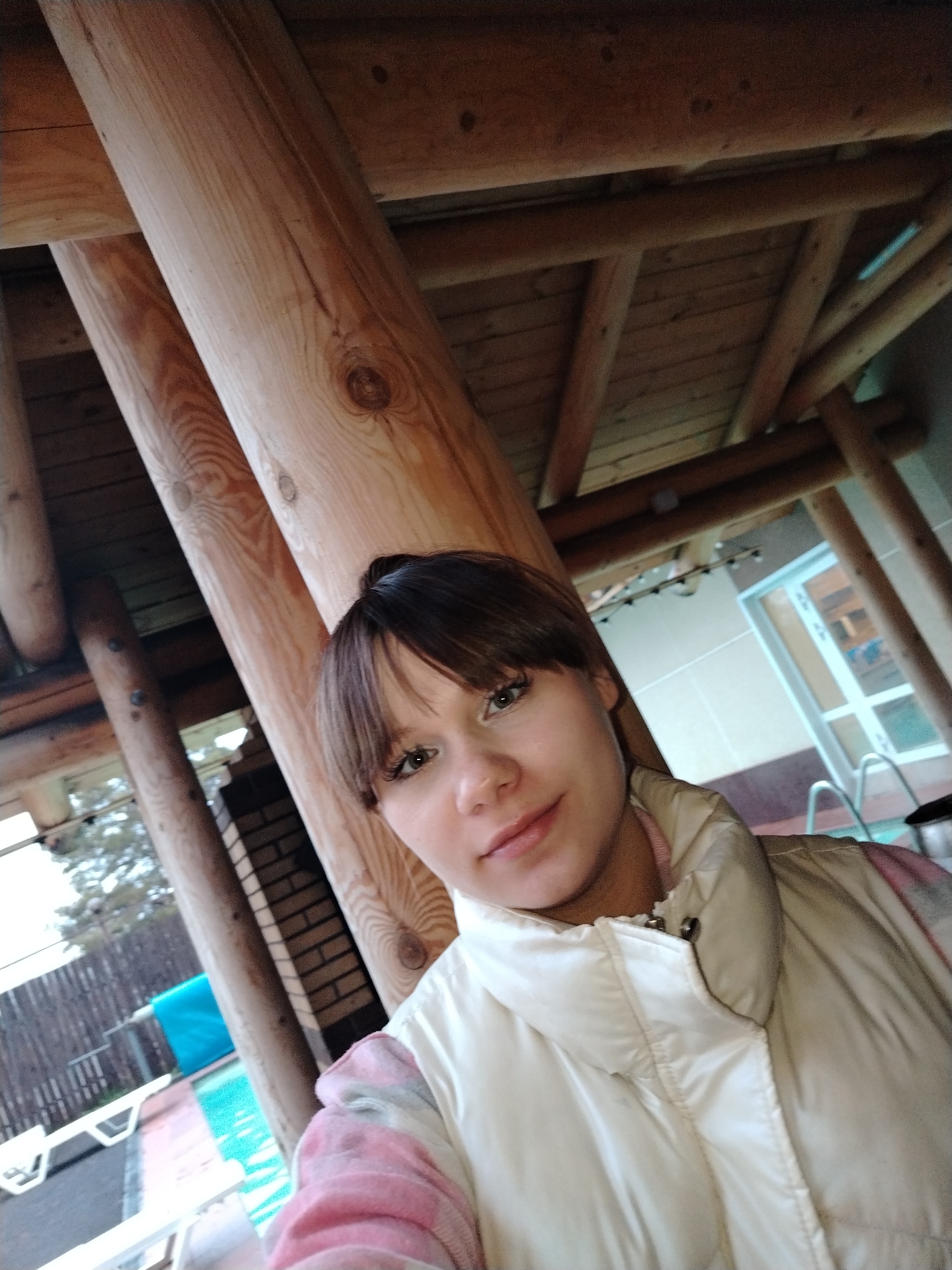 A young woman is featured in a selfie, looking directly at the camera with a slight, friendly smile. She has short, dark brown hair with bangs that frame her forehead and light-colored eyes. She is wearing a light beige or off-white puffy vest over what appears to be a pink and white patterned top or sweater.
The scene is set outdoors under a sturdy wooden structure, possibly a veranda, patio, or a part of a wooden house, characterized by prominent round and squared wooden beams and a planked wooden ceiling overhead. The lighting suggests it is daytime.
In the background to the left, beyond the wooden supports, a dark wooden fence, some green foliage, and an outdoor swimming pool are visible. The pool contains blue water and has white lounge chairs lined up beside it. A bright blue object is also partially seen near the pool. To the right, another section of a swimming pool is visible, along with a metal ladder leading into it. Behind her on the right, there is a light-colored wall with a large glass door or window, suggesting an adjacent building or indoor area, possibly a pool house. The overall setting implies a recreational area, possibly in a private residence or a leisure facility in Chaikovsky, Russia. There is no visible text in the image.