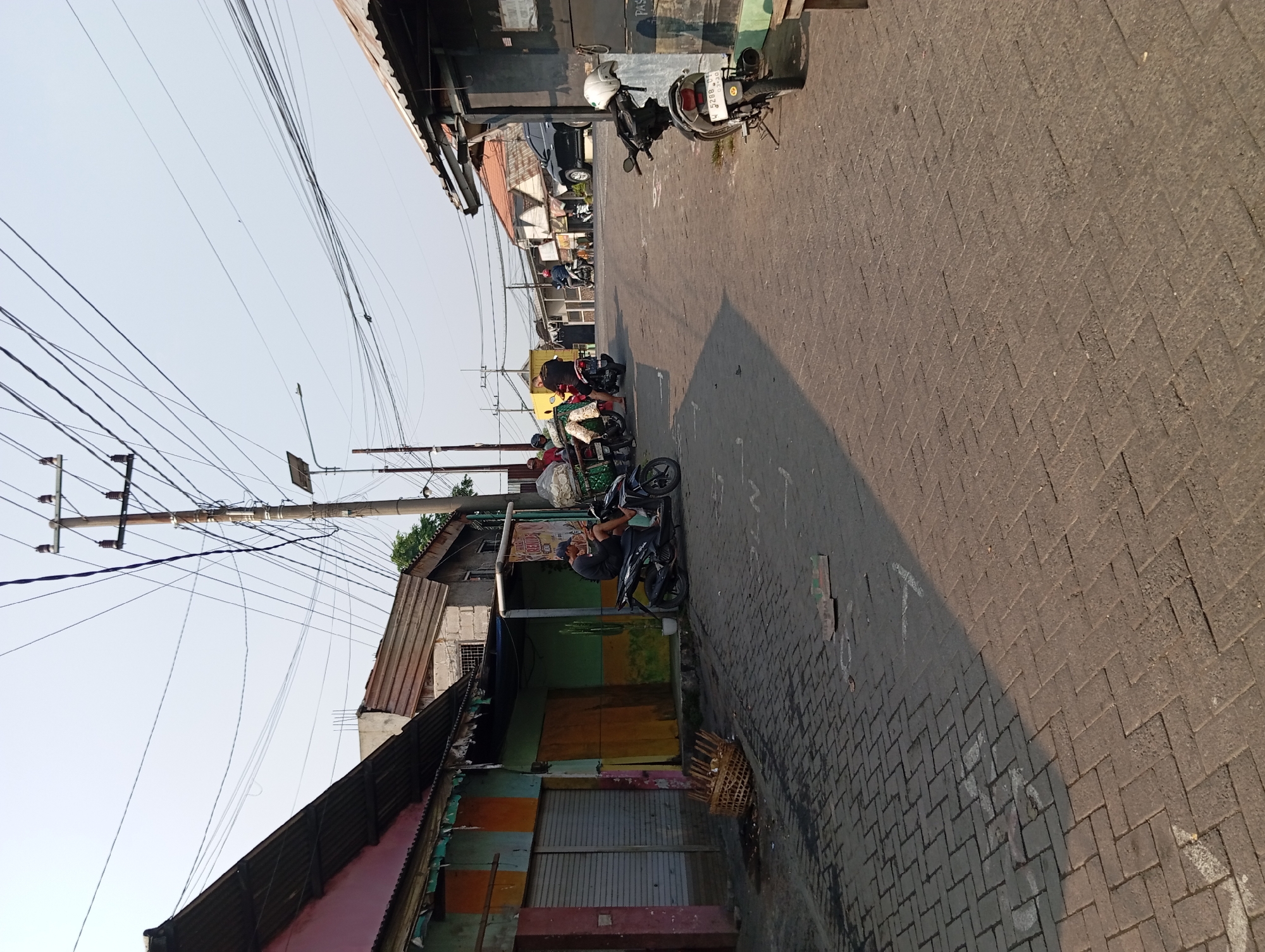 The image depicts a street scene in Kepulungan, Indonesia. The road is paved with brickwork and slopes slightly upwards into the distance. On the left side of the street are several buildings with corrugated metal roofs and painted facades in various colors, including pink, orange, green, and yellow. Some of these buildings appear to be shops with roller shutters. In front of one of these buildings, a man wearing a dark t-shirt and a blue cap is seated on a black motorcycle, looking at his phone. A woven basket filled with goods is placed on the ground near the motorcycle.
Several other motorcycles are parked or in use on the street. Further down the street, more buildings line both sides, some with more traditional tiled roofs. Power lines crisscross the sky above, supported by utility poles. In the background, a black SUV is visible on the road, and other vehicles and people are present, suggesting a moderately active urban environment. The sky is clear and pale blue, indicating daylight, and the lighting suggests the sun is either rising or setting, casting shadows on the road.