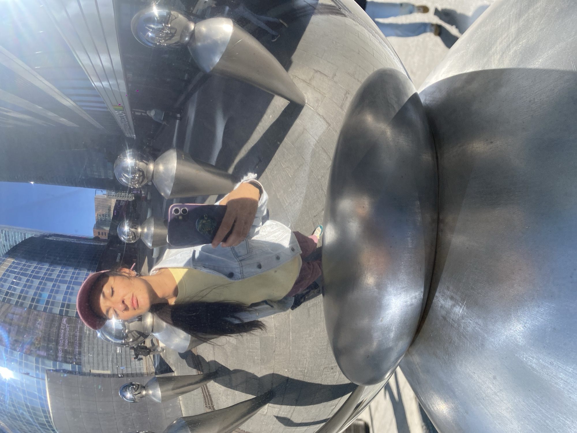 A young woman with dark hair and a maroon beret is taking a selfie in front of a large, reflective, metal sculpture in an urban area. She is wearing a yellow shirt and a light denim jacket. The sculpture reflects the surrounding buildings, including a tall glass skyscraper. The ground appears to be paved with light-colored stone or concrete. The specific location cannot be determined with certainty, but based on the provided description (city of Khimki, Russia), this information might be inaccurate since the buildings in the reflection appear to be American.