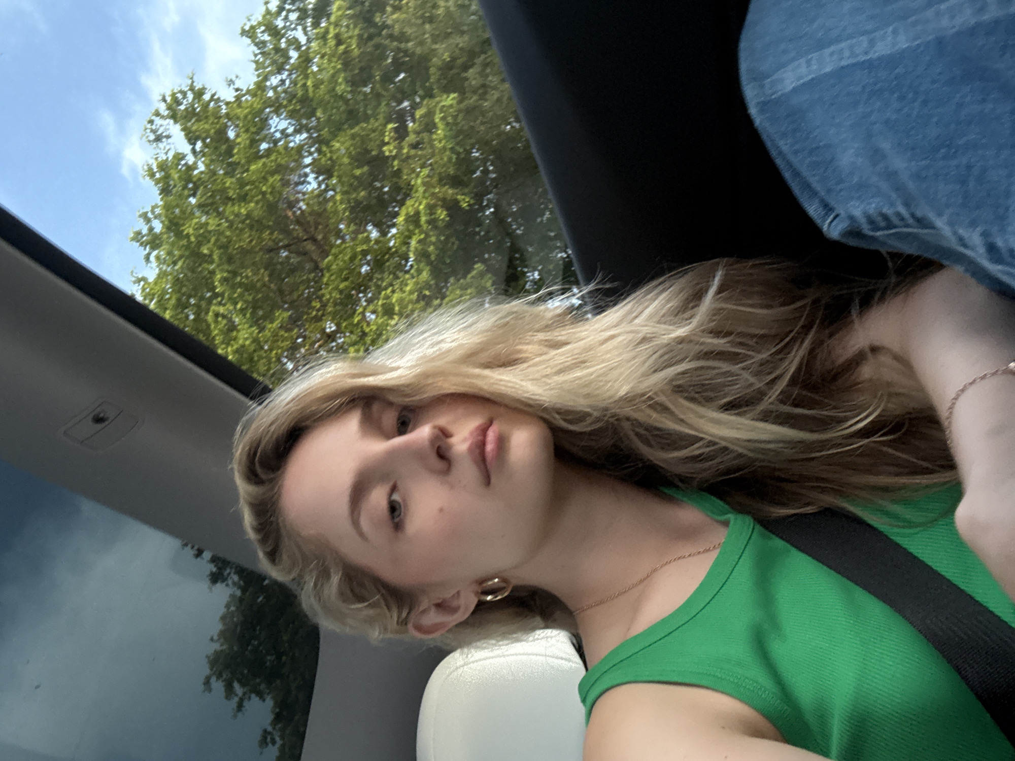 A young woman with blonde hair, wearing a green tank top and blue jeans, is seated in a vehicle. She has a necklace and hoop earrings. Through the car window, lush green trees and a blue sky with scattered clouds are visible. The car appears to be in motion, suggesting a travel scenario. The lighting suggests it is daytime. The scene is likely captured within Coral Gables, United States of America, given the vibrant foliage typical of the city's climate and aesthetic.