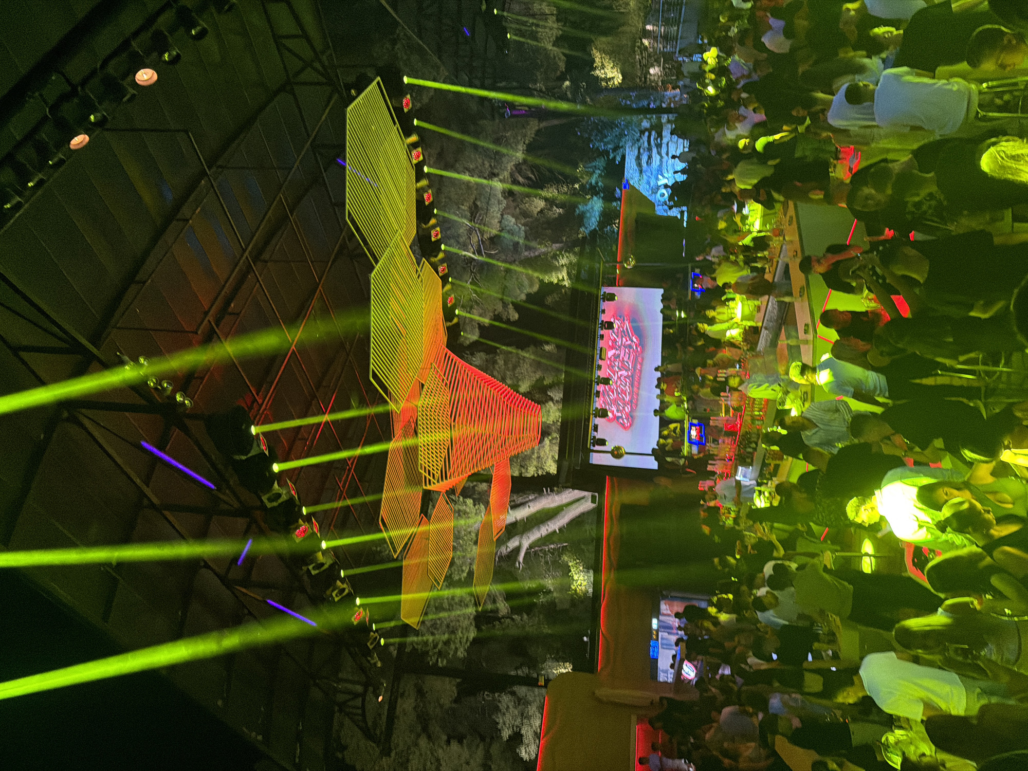 This image depicts a crowded nightclub scene in Kassandra Municipal Unit, Greece. A large group of people are visible, many of whom are standing and socializing. The environment is dimly lit, with vibrant green and purple spotlights illuminating the space. A large screen displays graphics, and various lighting installations, including hexagonal shapes, hang from the ceiling. The overall atmosphere appears to be lively and energetic.