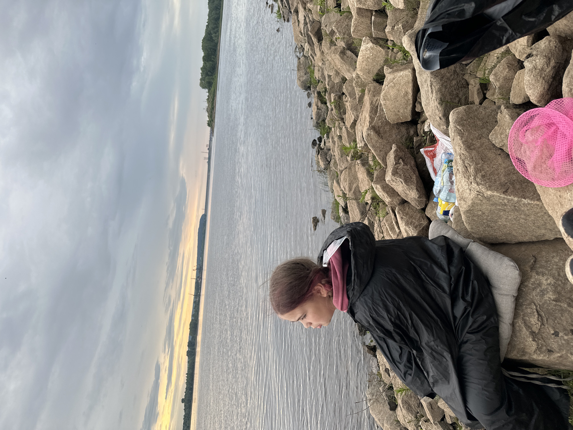 This media file depicts a young person sitting on a rocky shore next to a body of water. The person is wearing a black hooded jacket and grey leggings, and appears to be looking down at the water. The setting is a lake or river with a tree-lined bank visible in the distance. The sky is overcast with some hints of yellow and orange light, suggesting it may be sunrise or sunset. There are several rocks of varying sizes scattered along the shore. Among the rocks are a black plastic bag, some plastic bottles, and a pink mesh bag. The overall mood is calm and perhaps contemplative. No visible text is present in the image. The location context is Chaikovsky, Russia.