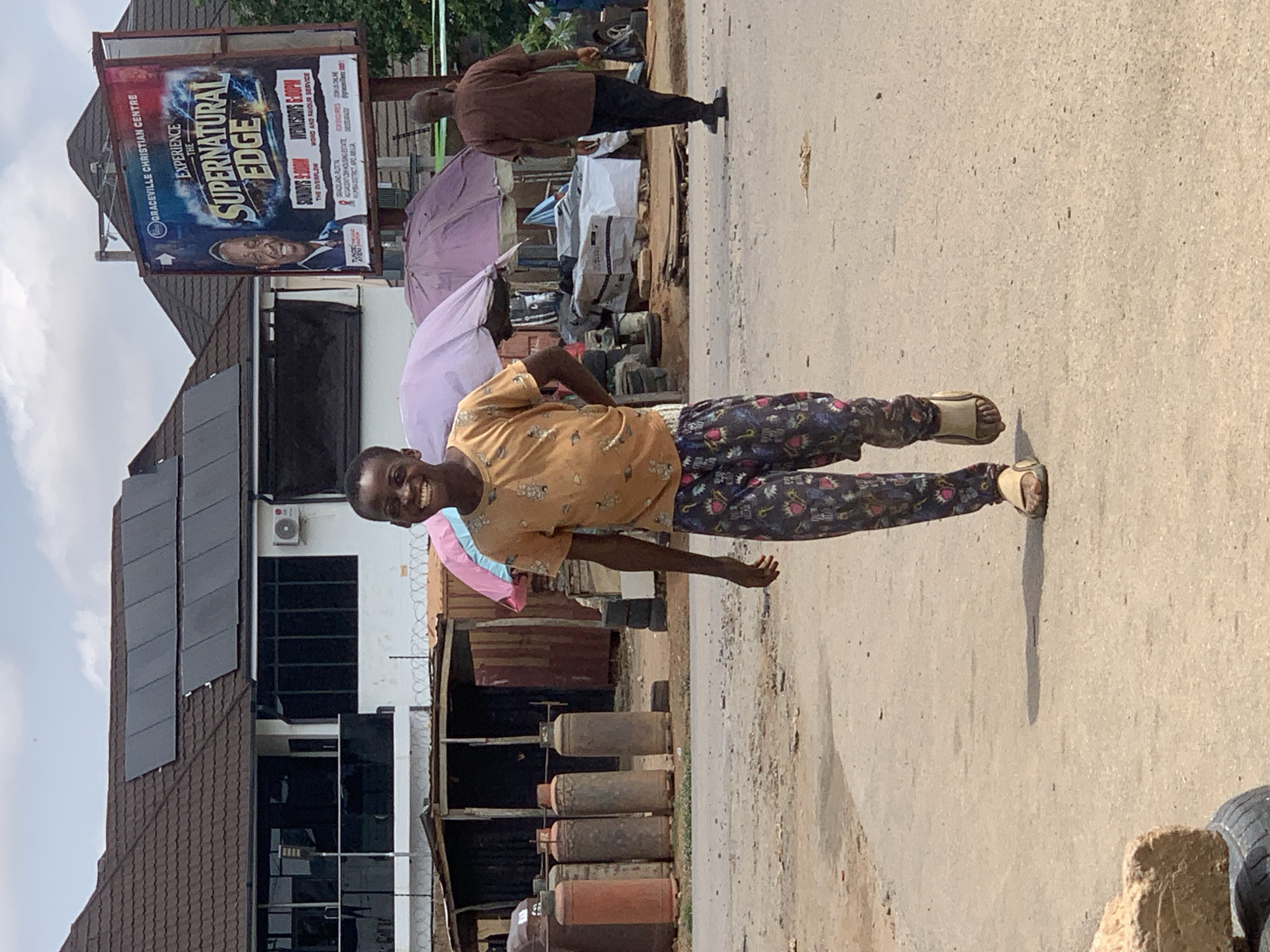 A young boy with a bright smile is walking towards the camera on a dusty, paved road. He is wearing a light orange t-shirt with a pattern of small white figures and patterned trousers with blue, red, and white designs. His feet are clad in light-colored sandals. The environment suggests a somewhat informal urban setting, possibly a market area, with stalls, gas cylinders, and tarpaulins visible in the background. A large billboard advertising a "Graceville Christian Centre" event titled "Experience The Supernatural Edge" is also present. Further in the background, another man is walking away from the camera. The sky is partly cloudy, indicating daylight. The location is the city of Abuja, Nigeria.