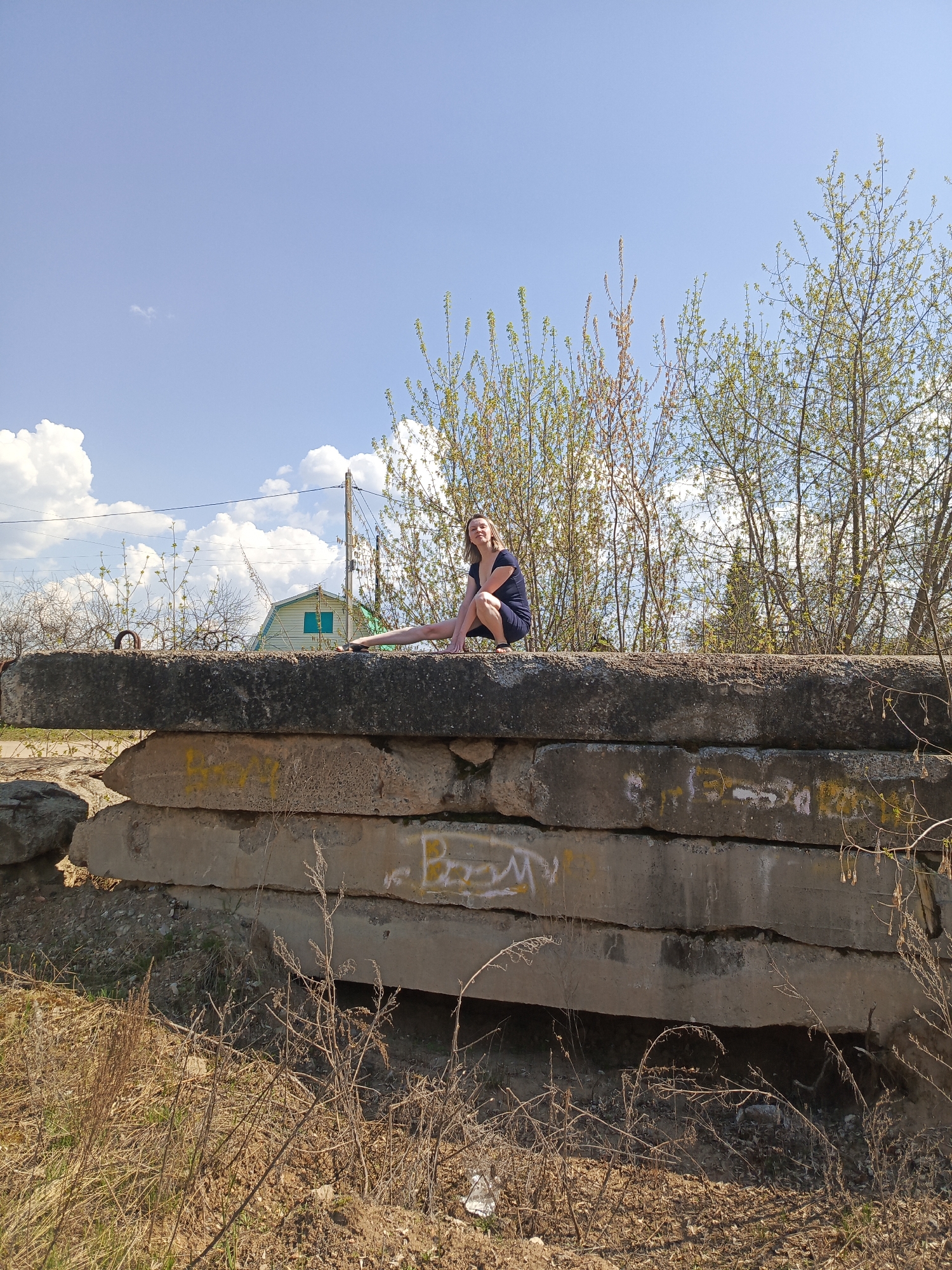 The image depicts a woman sitting atop a concrete structure in an outdoor setting. The structure consists of several large, stacked concrete slabs, some of which are covered in graffiti and yellow paint. The background includes trees with emerging green leaves, a bright blue sky with scattered clouds, and a building with a teal-colored roof. Dry grass and foliage are visible in the foreground. The location is identified as being in the city of Chaikovsky, Russia. The woman is wearing a dark dress and sandals, and she is positioned in the middle of the top slab. She is looking directly at the camera.