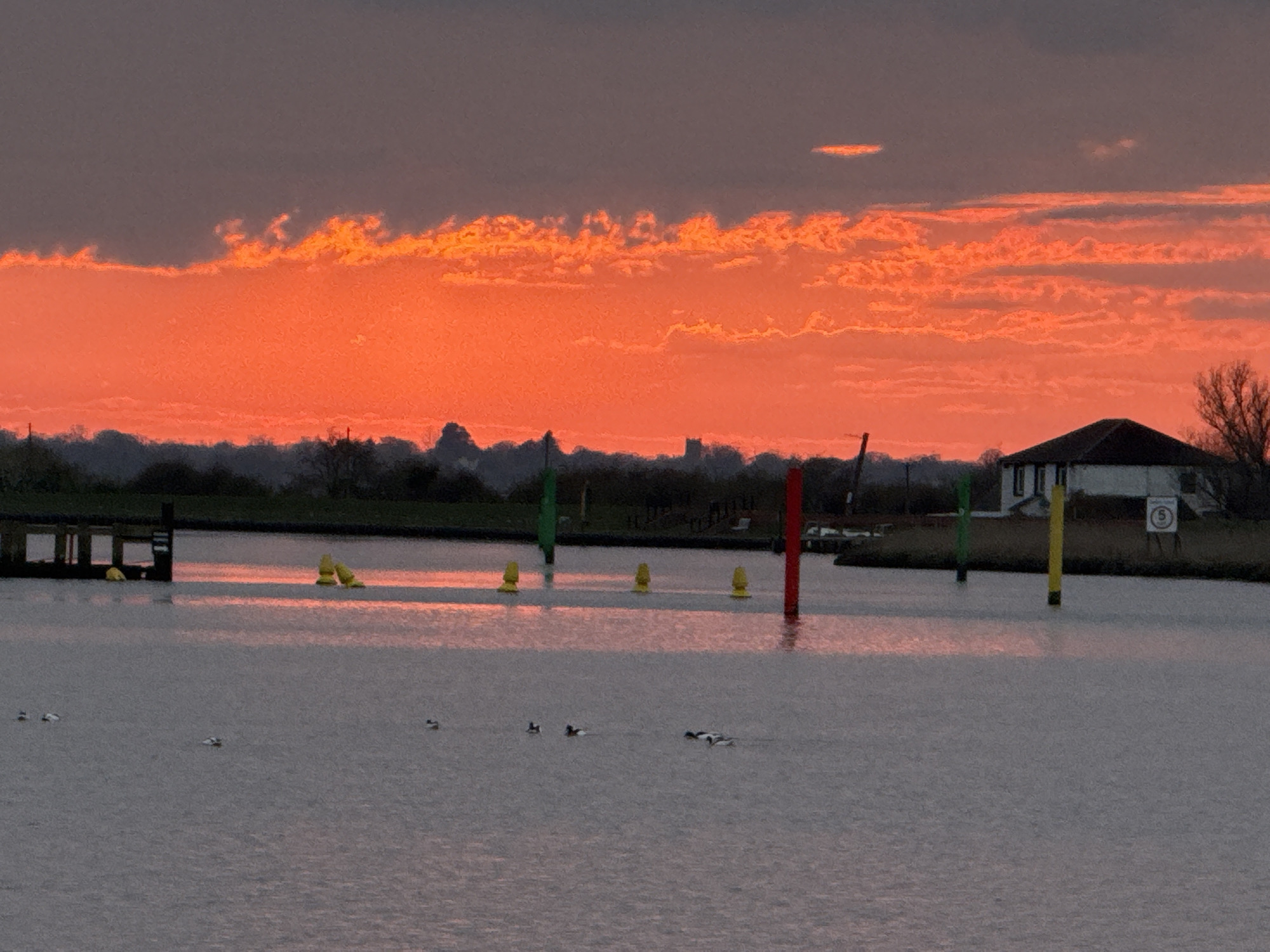 A landscape captures a water view at dusk or dawn, potentially the River Bure. A vibrant orange-red sky filled with wispy clouds dominates the upper portion of the frame, transitioning to a darker gray sky at the very top. Below, the water reflects the colors of the sky. In the middle ground, there's a dark silhouette of land with trees and indistinct structures, including a possible church spire. Several colorful poles – green, red, and yellow – stand in the water, possibly navigational markers, spaced along the frame. Small groups of waterfowl float on the water. On the right side of the horizon, a building with a dark roof is visible. A floating dock and some small buoys are on the left, near another dock. The scene suggests a location near the Norfolk Broads, possibly in or around Great Yarmouth, United Kingdom.