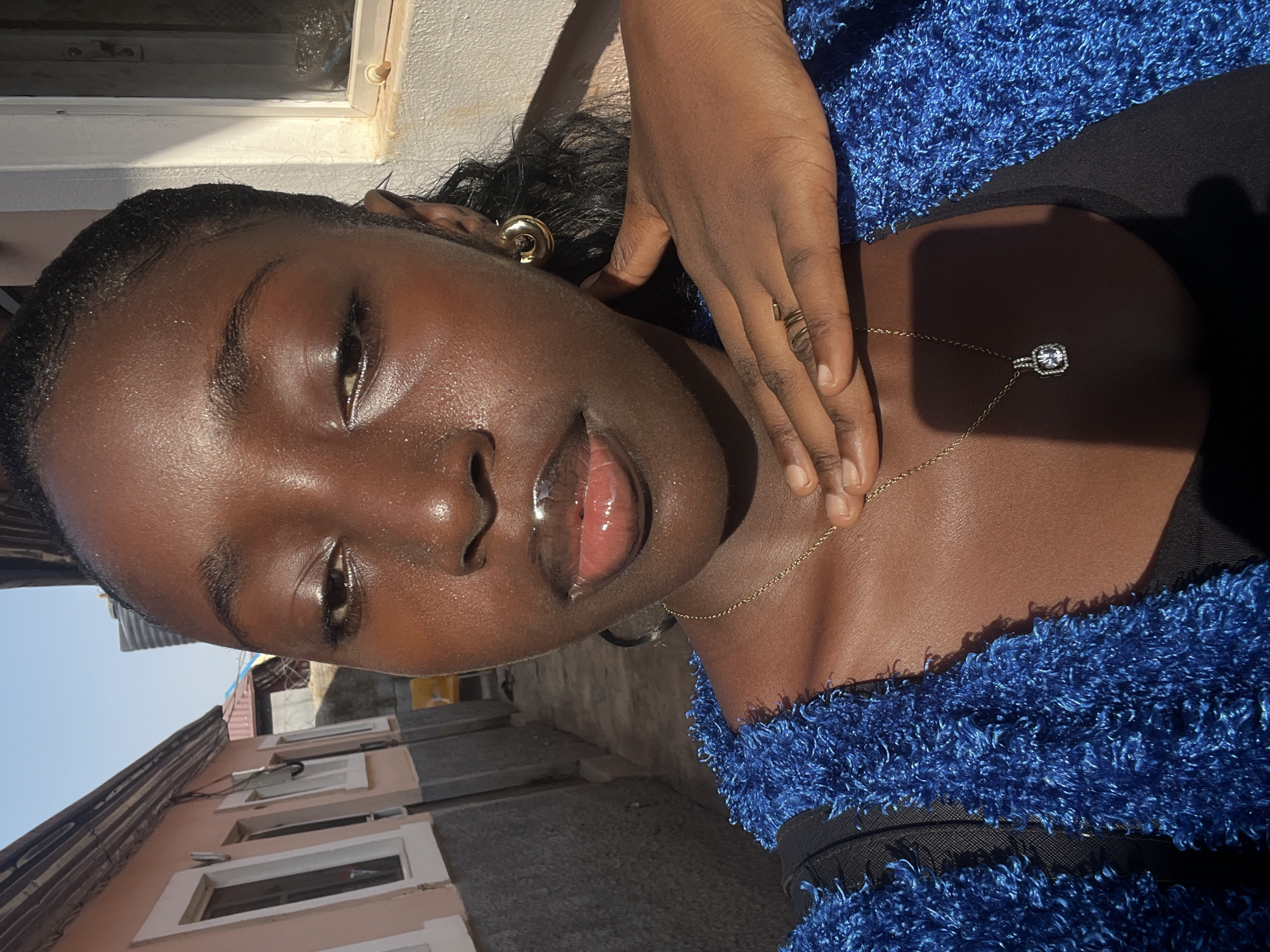 The image shows a young woman with dark skin and curly hair. She is wearing a blue, textured top over a black garment. A gold necklace with a pendant rests on her chest, and she has a gold ring on her finger. She is also wearing gold hoop earrings. Her skin appears to be glossy, possibly due to makeup or skincare products. Her hand is positioned near her neck.
In the blurred background, a building with light-colored walls and windows is visible. Based on the architectural style and the general environment, it can be inferred that the location is likely in a residential area of Abuja, Nigeria.