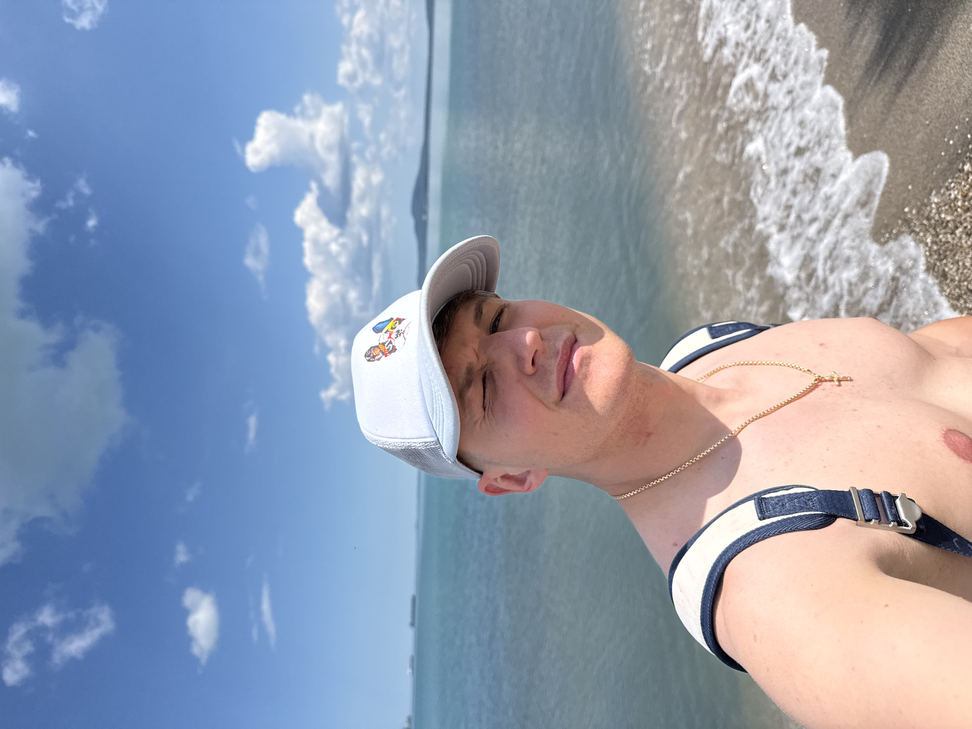 A medium shot depicts a man on a beach in Burgas, Bulgaria. The man is facing right with his head tilted to the left. He has fair skin, short hair, and is wearing a white cap with cartoon decals. A gold necklace with a cross pendant adorns his neck. He is wearing a blue and white strap. The background features a beach with gentle waves meeting the shore, a body of water, a shoreline in the distance, and a blue sky with scattered clouds. The image appears to be taken in bright sunlight.