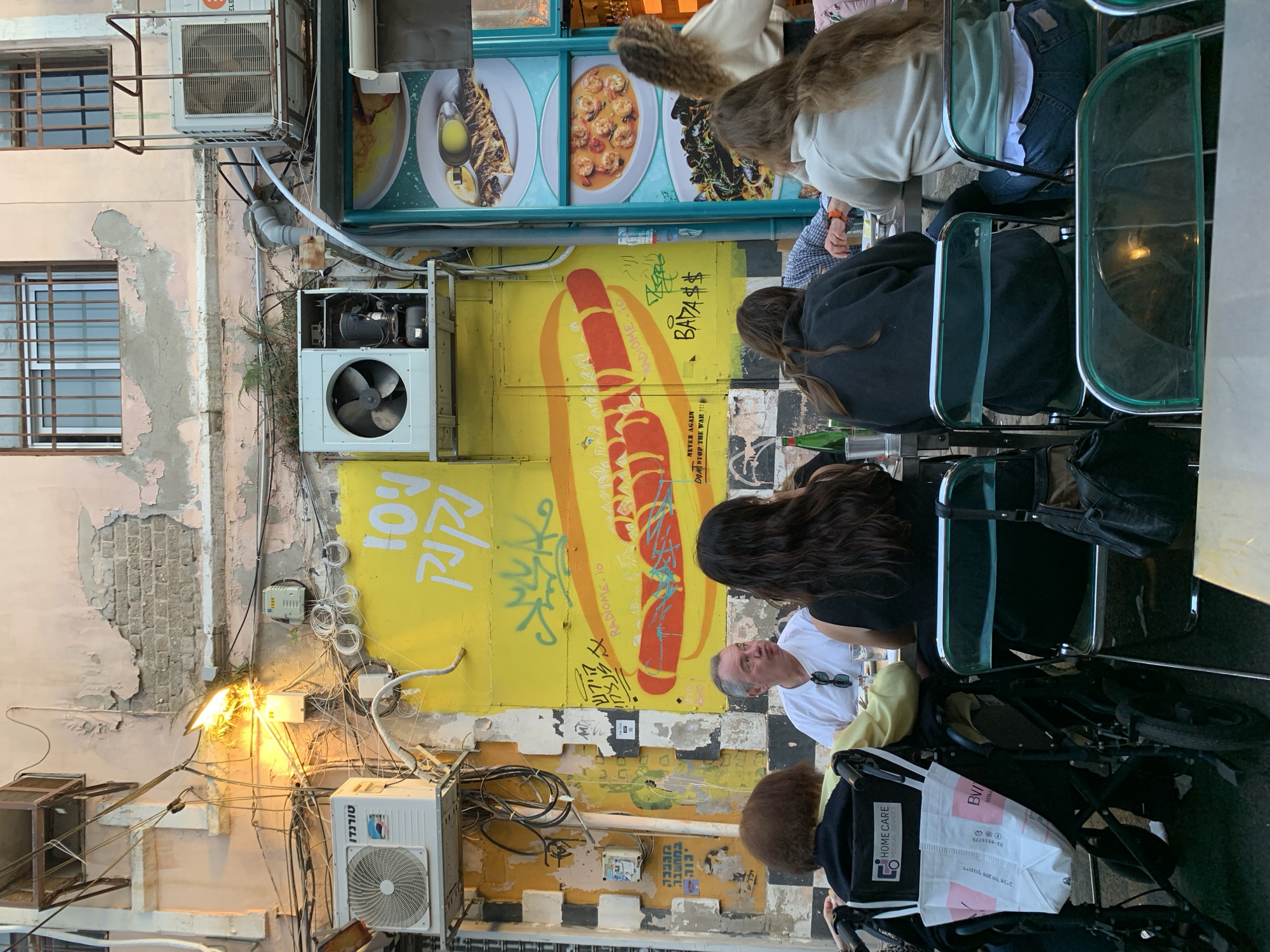 The scene is an outdoor dining area, likely in Tel Aviv, Israel, given the Hebrew graffiti and store names. People are sitting at tables. On the left, a weathered building features exposed layers of paint, multiple air conditioning units, and tangles of electrical wiring. An orange wall with a painted hotdog motif displays graffiti in Hebrew and English. A sign above the tables features images of food.
