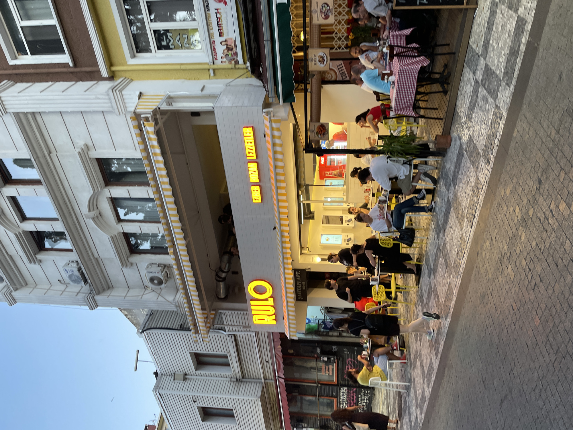 The image depicts a street-level view of a restaurant called "RULO" in Kadıköy, Turkey, bathed in the soft light of dusk. The restaurant has an awning with yellow and white stripes, and neon signs that read "RULO" and "EZHER BOZAN LEZZETLER." Several patrons are seated at tables both inside and outside the establishment.
Inside the restaurant, a red Coca-Cola refrigerator is visible. The outdoor seating area is set up on a patterned tiled ground, with a mix of white and bright yellow chairs around tables. Some tables are covered with red and white checkered tablecloths. People are actively engaged in dining and conversation, with waitstaff also present.
The architecture of the building above the restaurant is a mix of styles, with decorative pillars and multiple windows. Adjacent buildings are also visible, including one with a yellow facade and a sign that appears to advertise language courses. The overall atmosphere suggests a lively evening in a bustling urban environment.