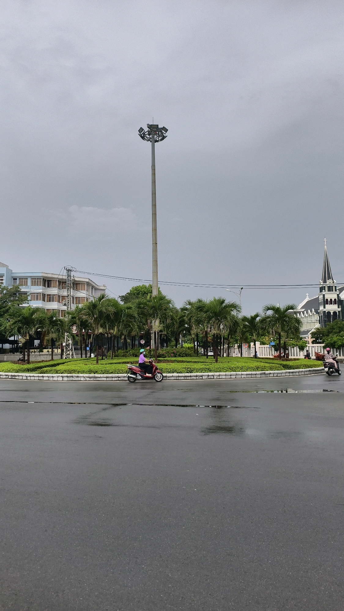 A wide shot shows a traffic circle with a tall pole light in the center. Palm trees and green landscaping are within the circle. To the left of the circle is a light blue building. To the right is a white and black church. A red scooter with a person in a purple shirt is on the circle. The ground is wet, indicating recent rain. The sky is overcast.