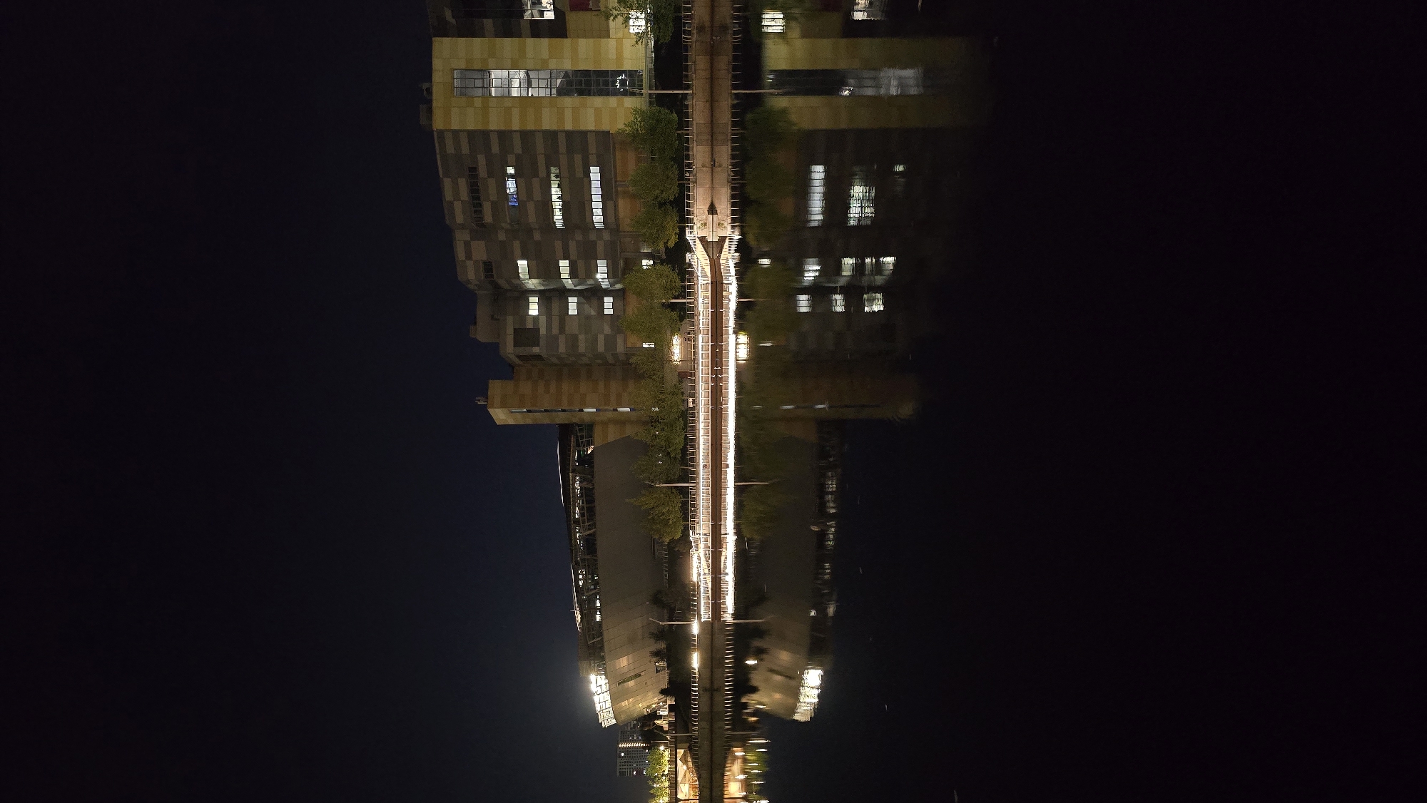 At night, a waterfront view showcases a modern, multi-story building with a striking facade composed of beige, grey, and yellow rectangular panels, interspersed with numerous lit windows. To the left of this building, a large stadium-like structure with a curved, illuminated roof is visible. The structures are reflected in the calm, dark water of a river or canal that occupies the lower half of the image. Trees line the bank between the buildings and the water. A pedestrian walkway with lights runs along the edge of the water. The sky above is dark and cloudless. The scene appears to be an urban setting, possibly a business or entertainment district, with the architecture suggesting a contemporary design. This location is identified as the area around the Obolon embankment in Kyiv, Ukraine.