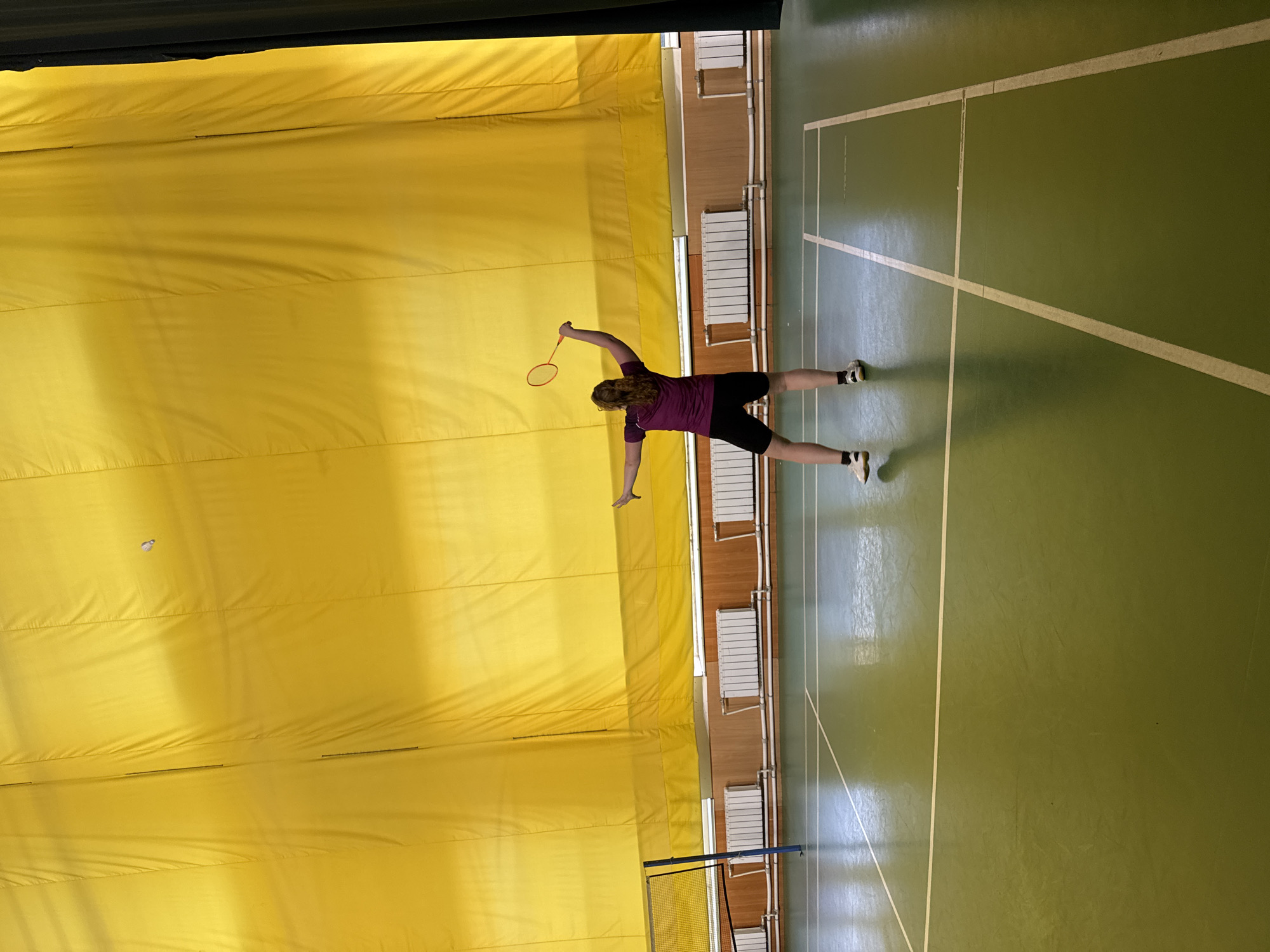 The image depicts an indoor badminton court within a gymnasium. A single individual, appearing to be an adult female with light brown hair pulled back, is observed from behind, positioned on the green, glossy court. She is wearing a purple short-sleeved top, black athletic shorts, and white athletic shoes with dark accents and white socks. Her right arm is raised and bent, holding an orange-framed badminton racket, indicating she is in the act of swinging or having just swung. Her left arm is extended outwards for balance. A white shuttlecock is visible in mid-air, slightly above and to the left of her racket, against the yellow background.
The court floor is a light green with clear white demarcation lines. To the left, a blue pole supports a white badminton net. The background features a large, bright yellow fabric or curtain covering the upper half of the wall. Below this, there is a section of light brown wainscoting or wood paneling. Further down, multiple white radiator units with horizontal white piping are mounted on the wall. On the far right edge of the frame, a dark, possibly black, curtain or wall section is partially visible. The specific location within the city of Saint Petersburg, Russia, cannot be determined from the visual information in the image.