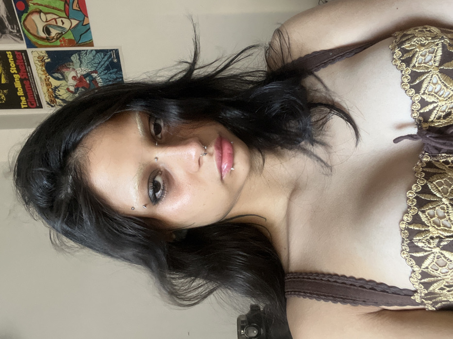 A woman with dark hair, light eyes, and pierced eyebrows, nose, and lip. She's wearing a brown lace top. Behind her on the wall are several posters, including one of Spider-Man and one with the words "The Rolling Stones Gimme Shelter." The image is rotated counterclockwise.