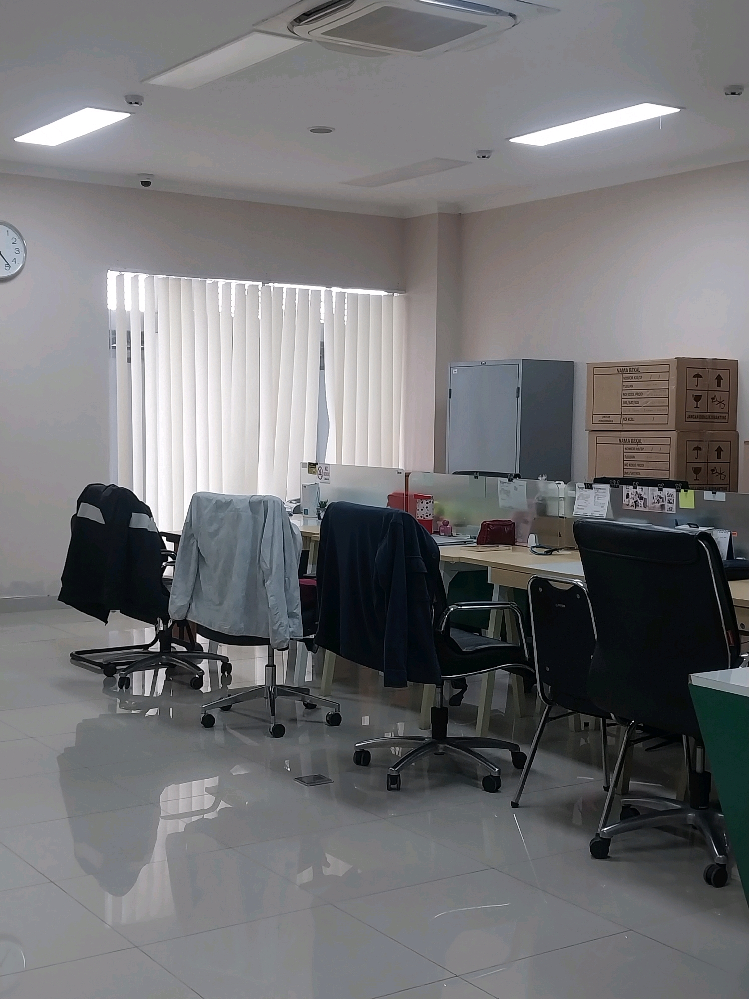 The image depicts an office environment in Sleman, Indonesia, possibly a co-working space or a small business office. The room features a row of desks with office chairs, some of which have jackets draped over them. The desks are partitioned by low-height dividers. In the background, there are stacked cardboard boxes, a filing cabinet, and vertical blinds covering a window. Overhead, fluorescent lighting panels and a ceiling fan are visible. A clock on the wall indicates the time. The floor is tiled and reflective, showing the reflections of the furniture and blinds. There are no people visible in the image. The overall impression is one of a functional, though sparsely populated, workspace.