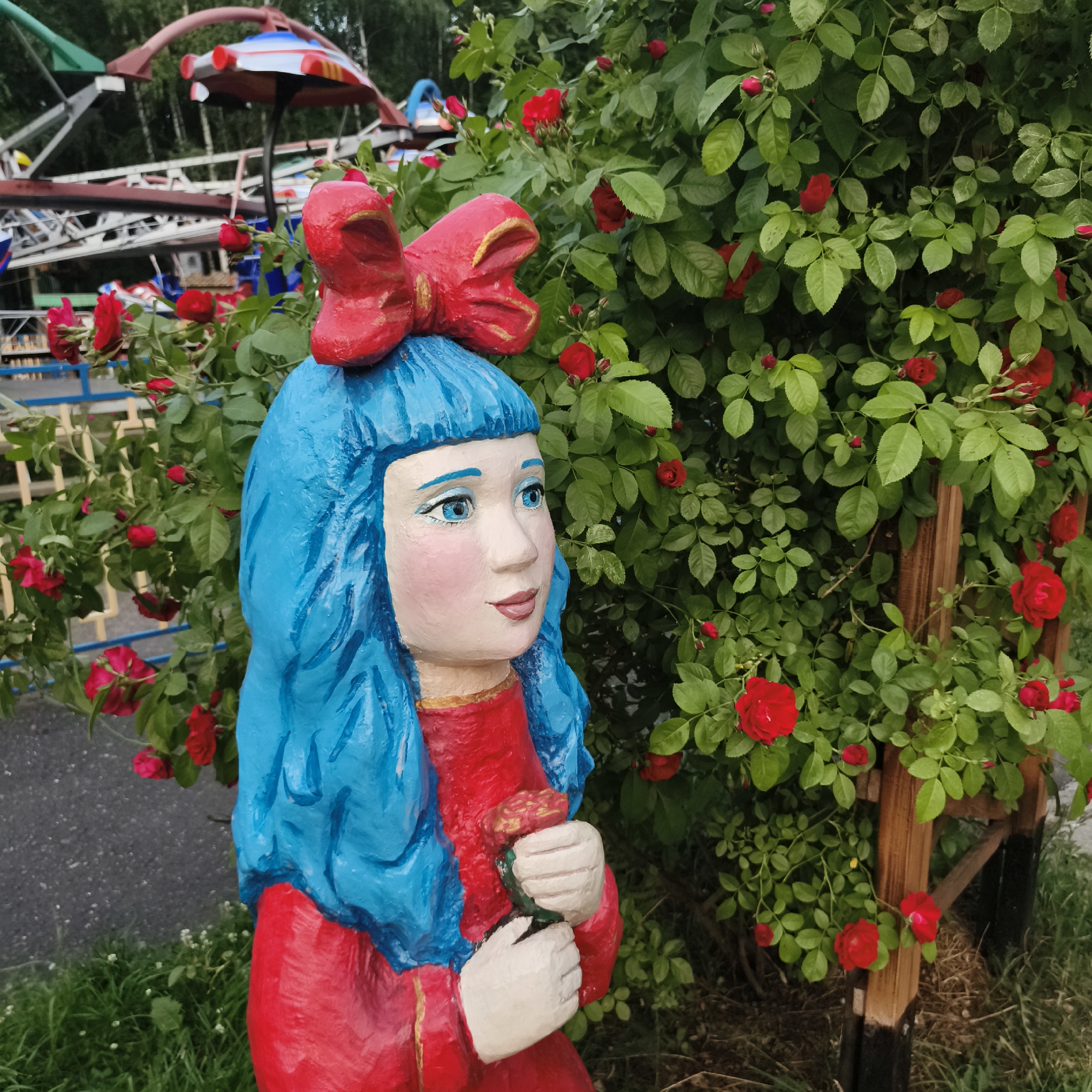 An outdoor, medium-shot, eye-level photograph captures a painted wooden sculpture of a young girl with bright blue hair and a red bow, wearing a red dress, standing in front of a rose bush with red roses. The girl is holding a pink object in her hands. The background features a children's amusement park ride, a wooden fence, and trees. The scene appears to be in a park or amusement area in the city of Chaikovsky, Russia.