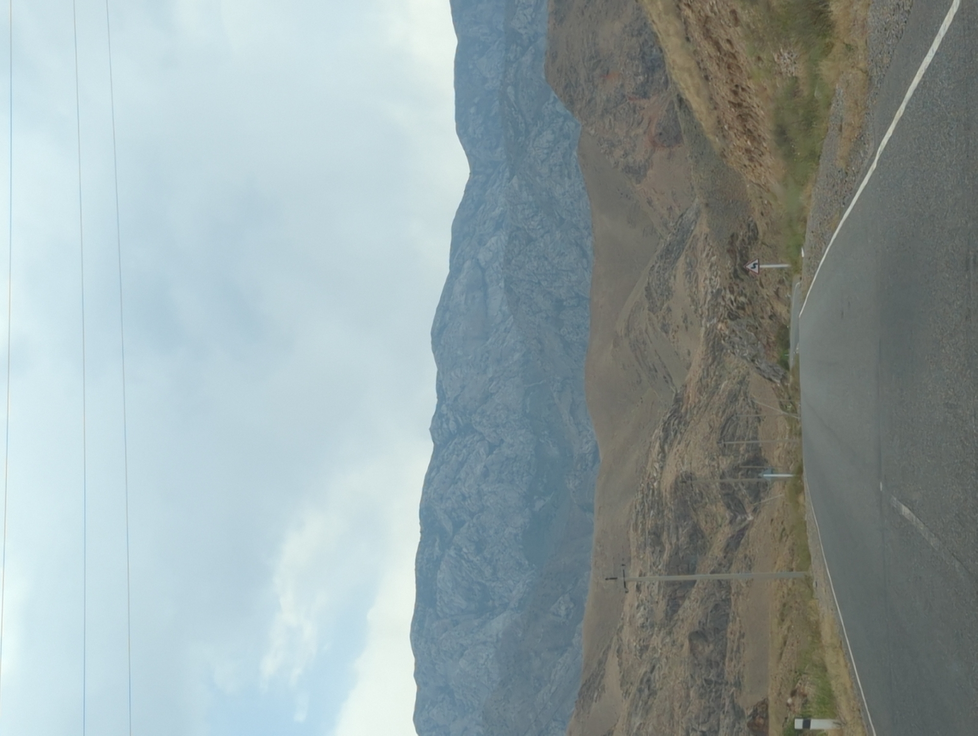 This is a photograph of a paved road in a mountainous, arid region. The road curves to the right as it recedes into the distance. On either side of the road are dry, grassy slopes with rocky outcrops. In the background, a large mountain range with jagged peaks is visible under a hazy sky. A few utility poles and a road sign are present along the roadside. The scene appears to be a rural or remote area, possibly a highway passing through a mountainous pass. The location is identified as Samarkandyk, Kyrgyzstan. There are no people or animals visible in the image.