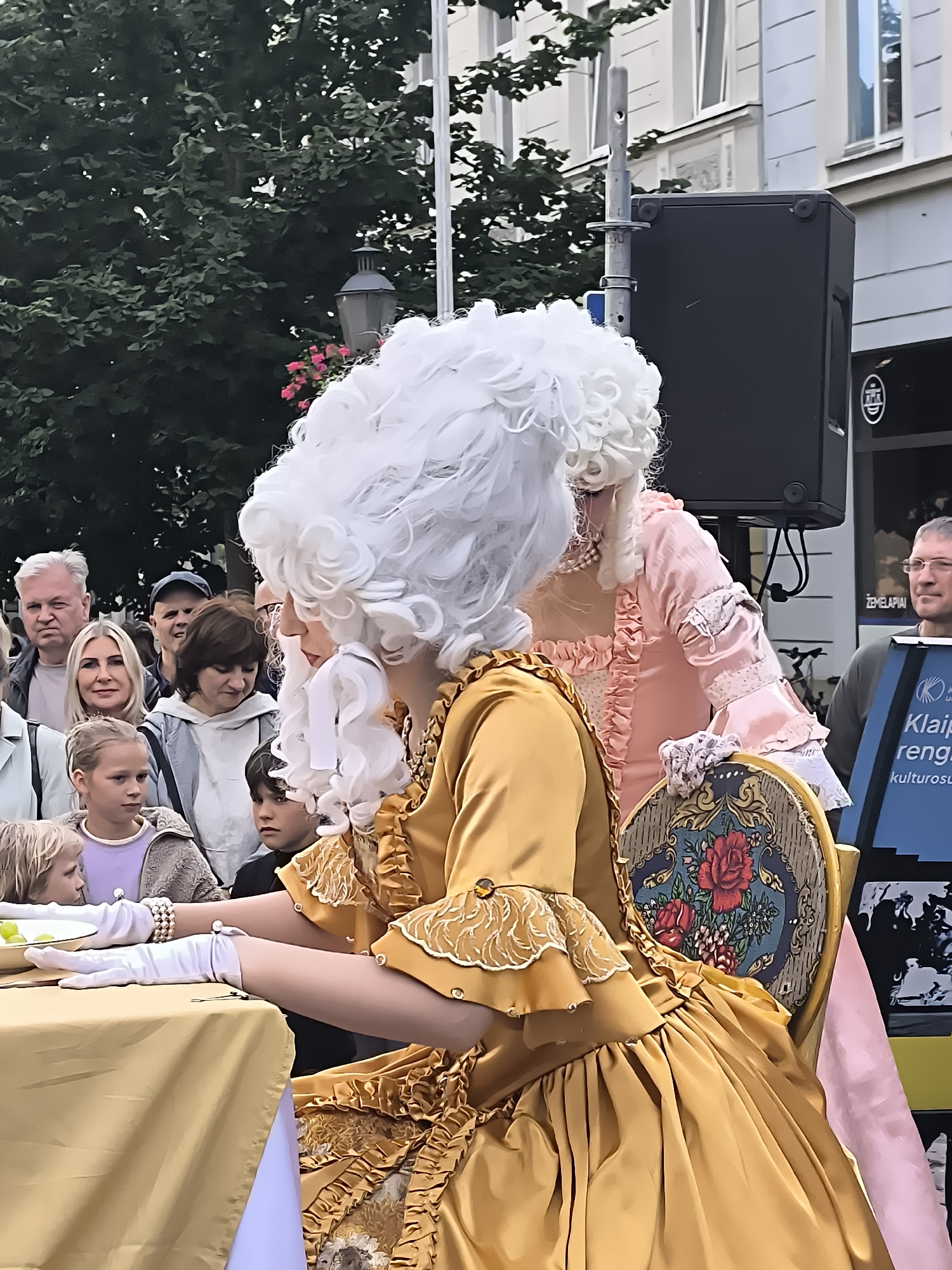 In Klaipėda, Lithuania, a historical reenactment event is underway. The main focus is a woman dressed in a lavish, 18th-century style golden gown, complete with a voluminous white wig and white gloves. She is seated at a table covered with a golden tablecloth, and her hands are resting on the table, suggesting she might be interacting with objects or performing a task. Beside her, another individual in a pink period costume and a similar wig stands. A crowd of spectators, including children and adults, are gathered behind them, observing the spectacle. In the background, a large black speaker is visible, indicating a public performance or presentation. The presence of trees and buildings suggests an outdoor urban setting.