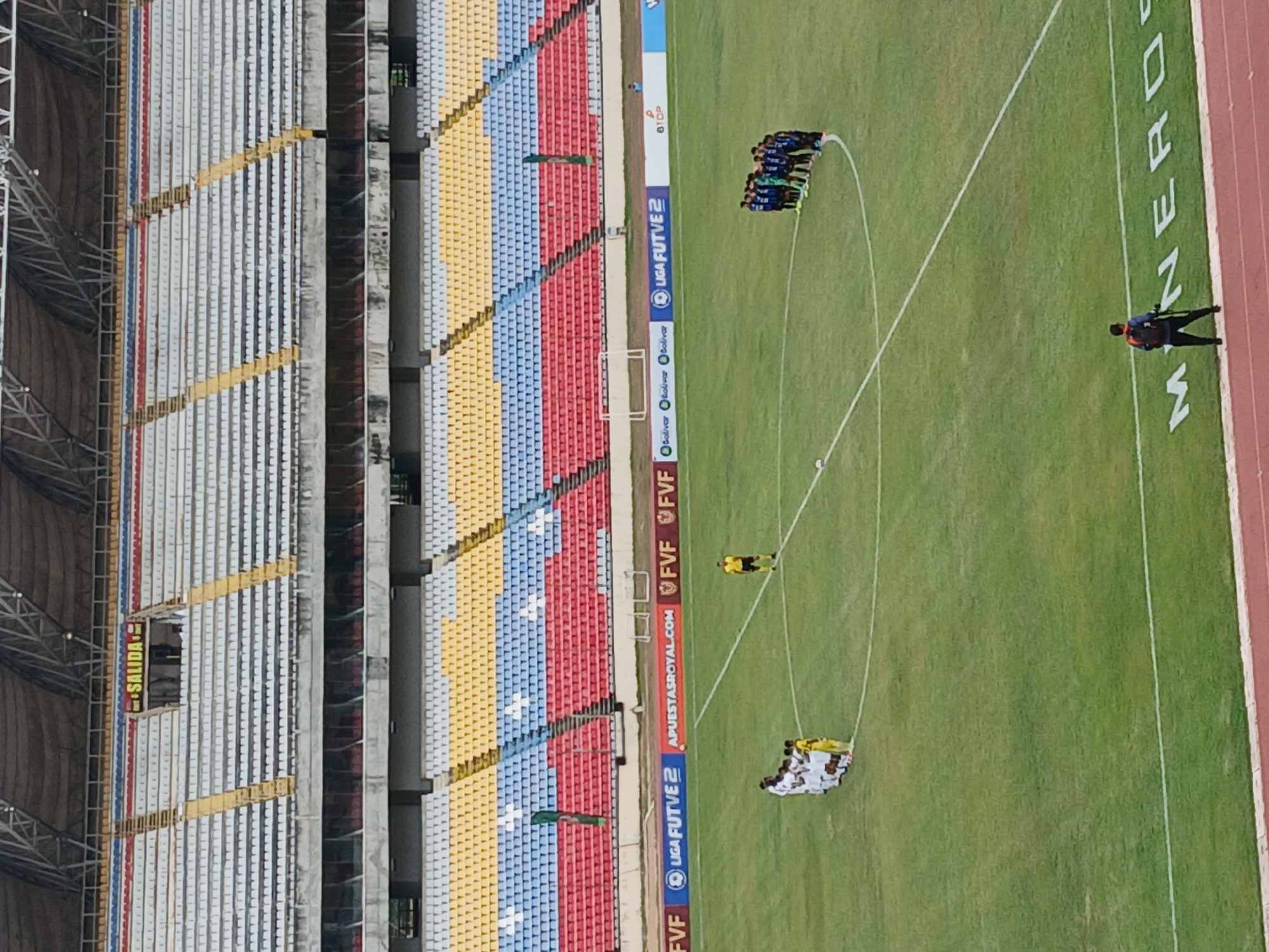 The image shows a soccer game being held in a large stadium with empty seats colored with white, yellow, blue, and red. The stadium has a metal frame and a yellow "SALIDA" sign is visible on the side. The playing field is green with white markings. There are two groups of players huddled together in the middle of the field. One group is wearing blue jerseys, while the other group is wearing white jerseys. One referee is positioned between the two groups of players. A person in a red and black jacket is standing near the sidelines close to the stadium's logo "MINERD". The location is Unknown, Unknown.