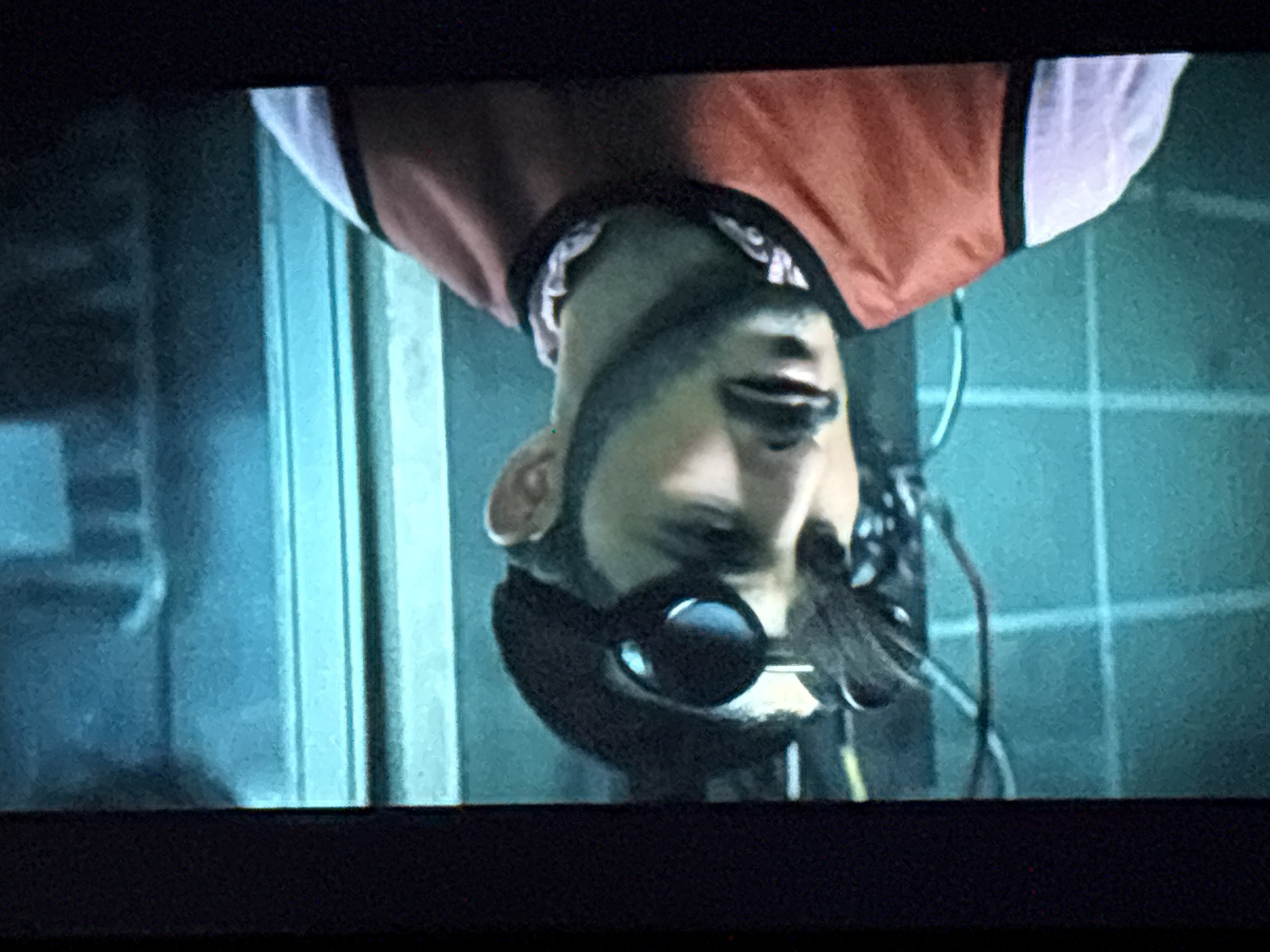 A man wearing a black beret and goggles is shown in a close-up shot. He has a beard and mustache and is wearing a pink shirt and an orange vest. The background is a teal-colored wall with some wires and a blue vertical panel. The image appears to be a still from a video or movie. There is no indication of the city of Dnipro, Ukraine.
