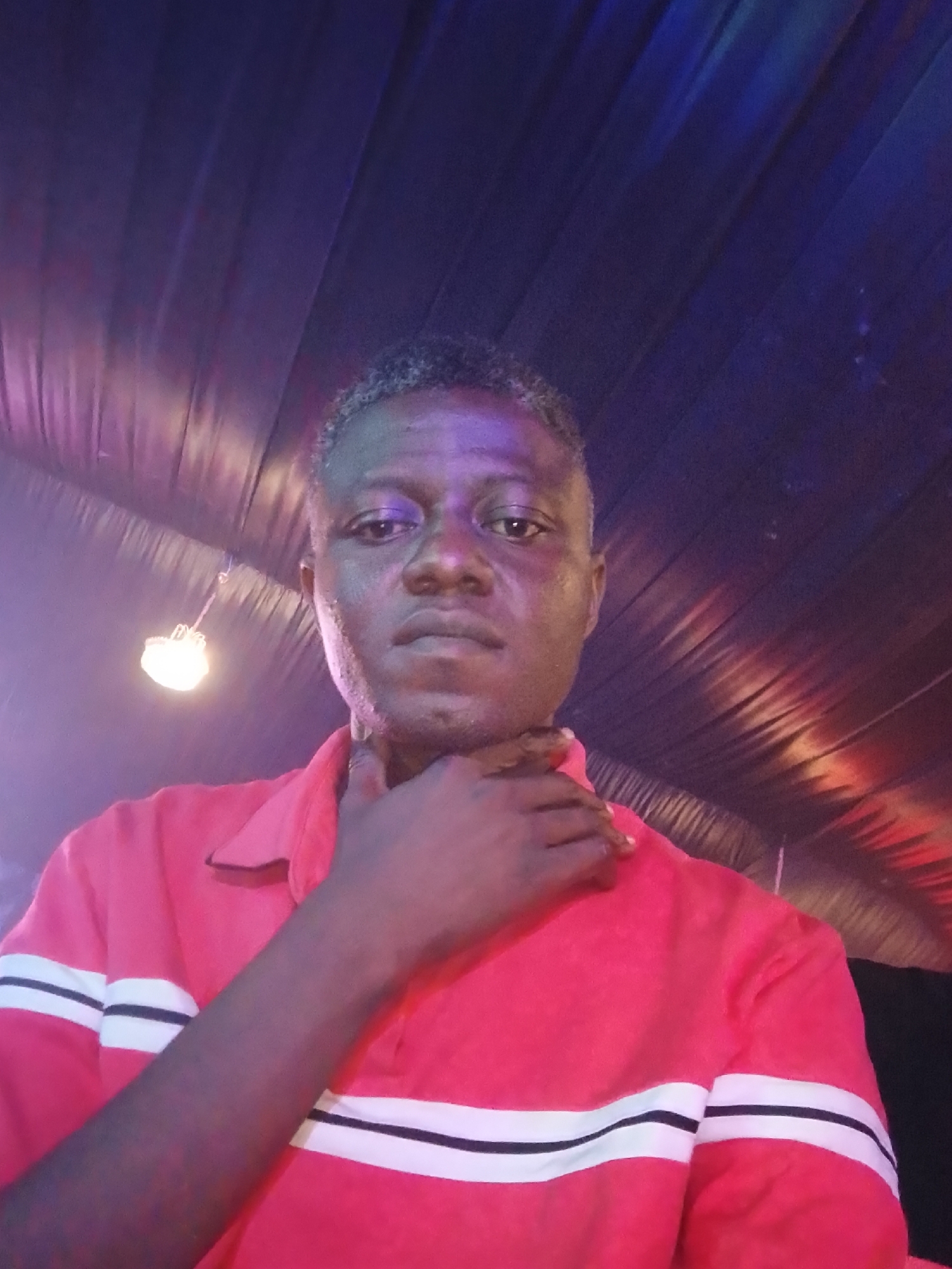 A selfie of a man is taken under uneven lighting. The man has short, graying hair and is wearing a red polo shirt with white stripes on the sleeves. He is holding his hand to his throat. Overhead, there is a fabric ceiling lit with mixed lighting including red and blue hues. The location is the city of Rido, Nigeria.