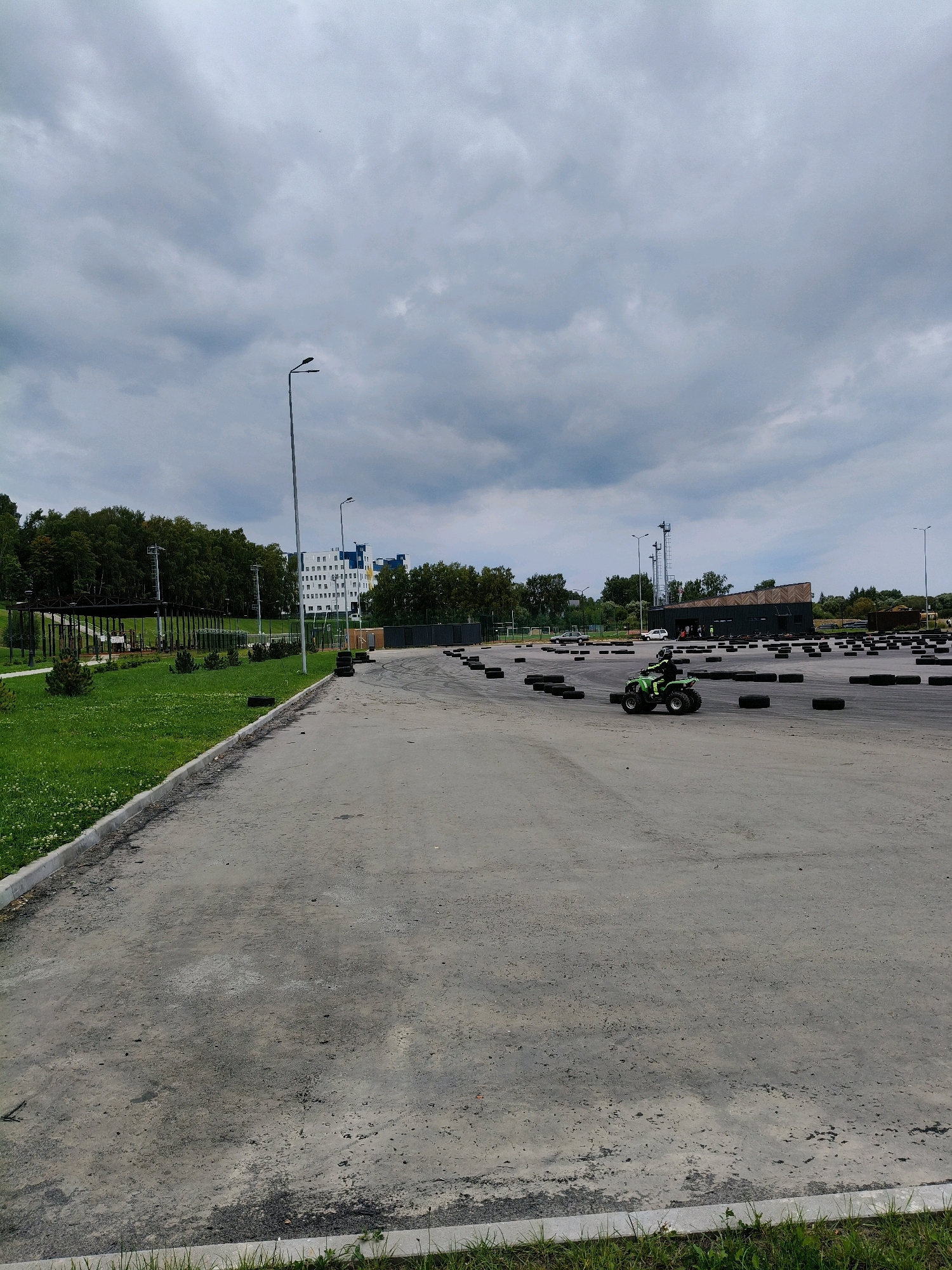 The image depicts an outdoor go-kart track located in Naro-Fominsk, Russia. The track is characterized by a grey asphalt surface and is bordered by numerous used tires, serving as markers or barriers. In the middle ground, a person wearing a helmet and dark clothing is operating a green four-wheel ATV. In the background, several multi-story buildings, a cluster of trees, and streetlights are visible under a cloudy sky. A grassy area with a paved walkway is situated to the left of the track. The overall scene suggests a recreational or sporting venue.