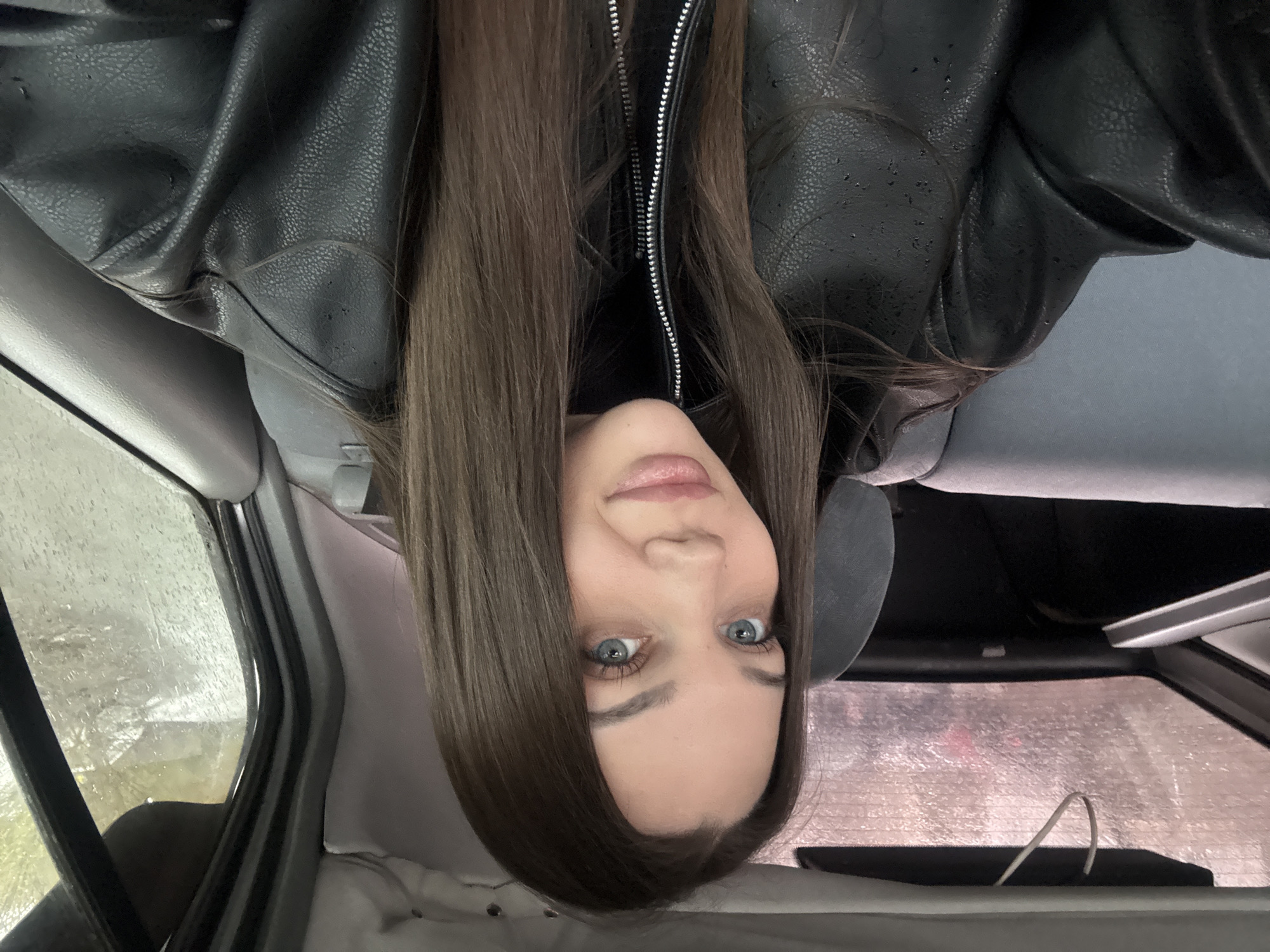 A young woman with long brown hair and blue eyes is inside a car. She is wearing a black leather jacket and a black turtleneck. The car appears to be stationary, with rain or condensation visible on the windows. The location appears to be Yekaterinburg, Russia; however, there's no direct evidence of that in the image.
