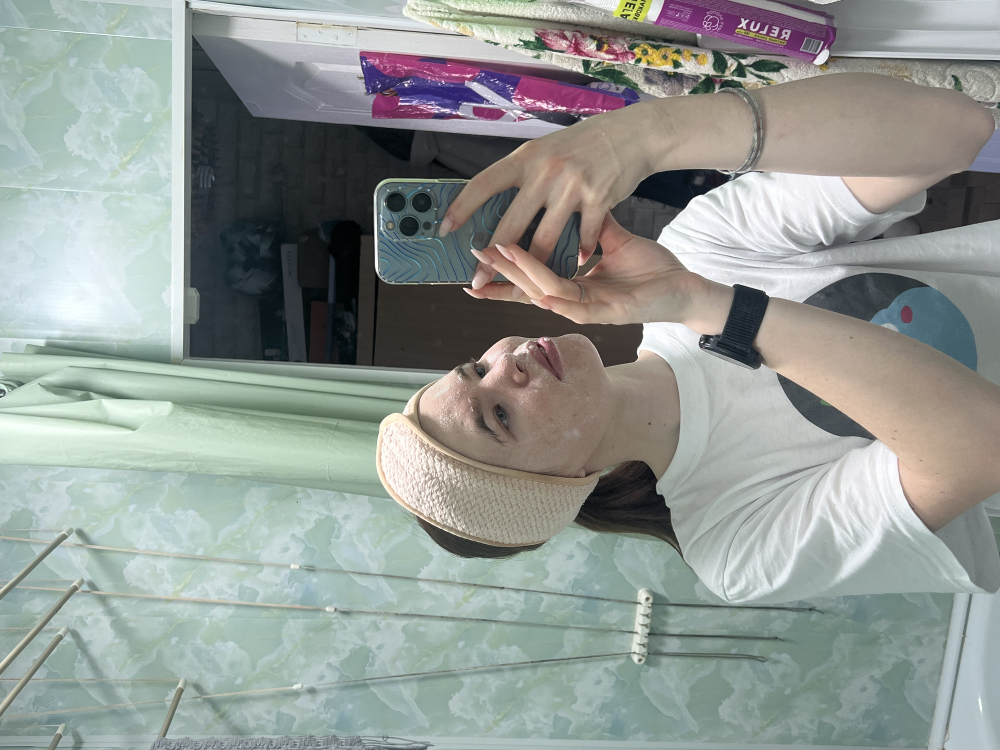 The image shows a person in what appears to be a bathroom. The person is fair-skinned, wearing a white t-shirt and a cream-colored headband. They are holding a smartphone, seemingly taking a selfie. Part of their face is visible with what looks like a facial mask. The background shows a bathroom with light green patterned tiles on the wall. There is a clothes drying rack in the lower left corner. There is a mirror with items on a shelf above it. The specific location within the city of Chaikovsky, Russia cannot be determined from the image.