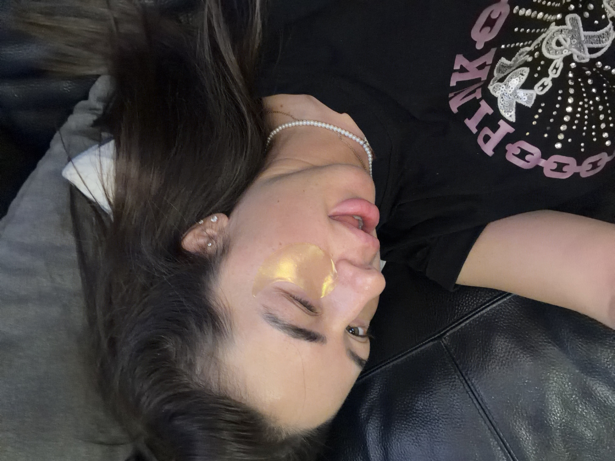 A woman is lying down on a black couch, facing sideways, with a pillow behind her head. She is wearing a black t-shirt with pink lettering and a design of chains and butterflies, adorned with rhinestones. She has on a pearl necklace and a gold under-eye mask on her right eye. Her left eye is partially closed, and her mouth is slightly open. Her dark brown hair is pulled back. The scene is indoors, with the couch dominating the foreground. There are no visible elements that indicate a specific location in Novi Iskar, Bulgaria.