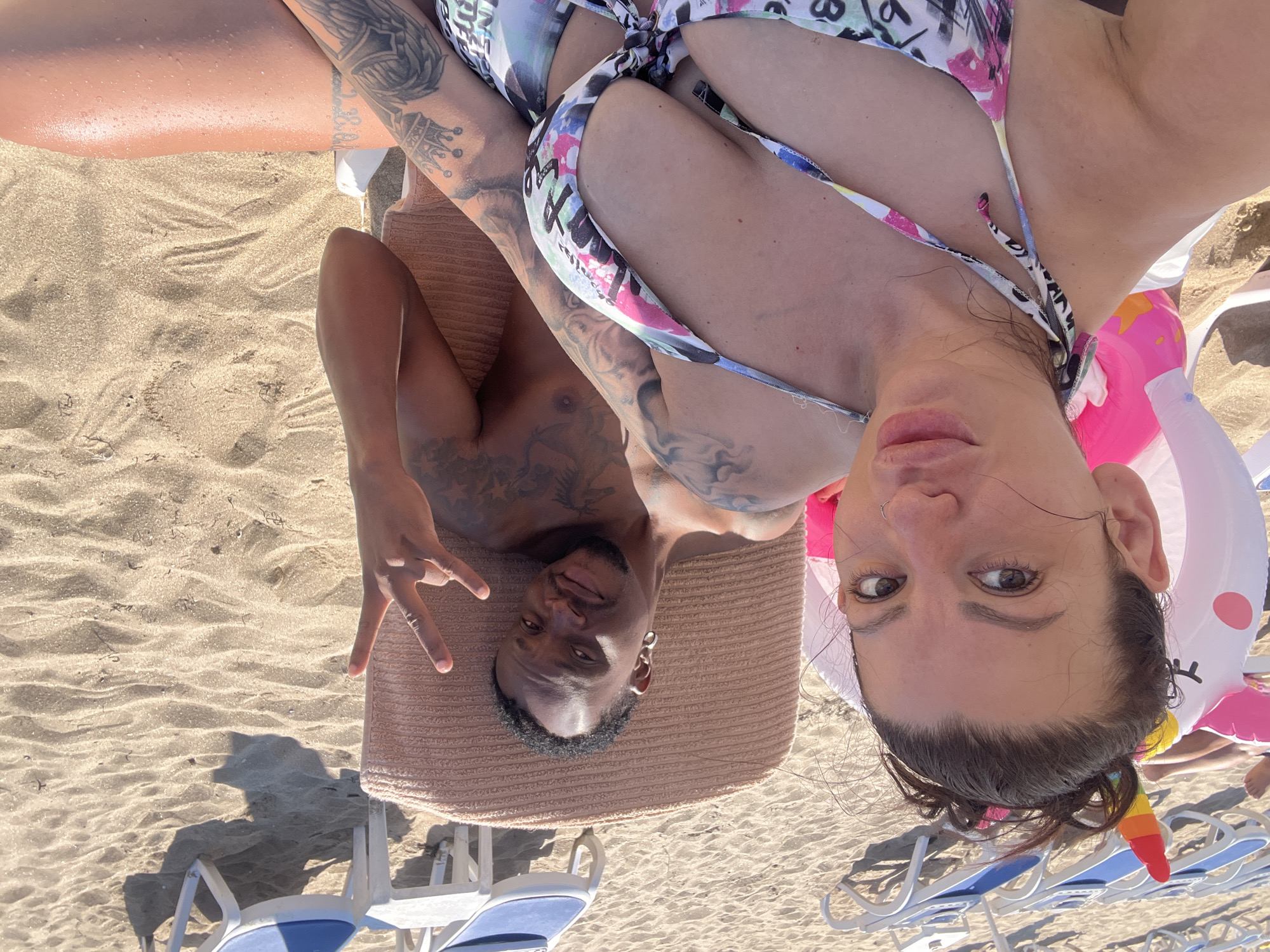 A medium shot captures a couple relaxing on beach chairs on a sandy beach. The woman, in the foreground, is taking a selfie, her face filling the left side of the frame. She has dark hair pulled up and is wearing a colorful bikini top. Next to her, a man is lounging on a recliner, turned slightly towards the camera and making a peace sign with his right hand. He has tattoos on his arms and chest. Behind them, several empty beach chairs are lined up, casting shadows on the sand. An inflatable unicorn float is visible behind the woman. The scene appears to be set on a sunny day at a beach, likely in Sozopol, Bulgaria, given the typical beach setups and architecture visible in the background.