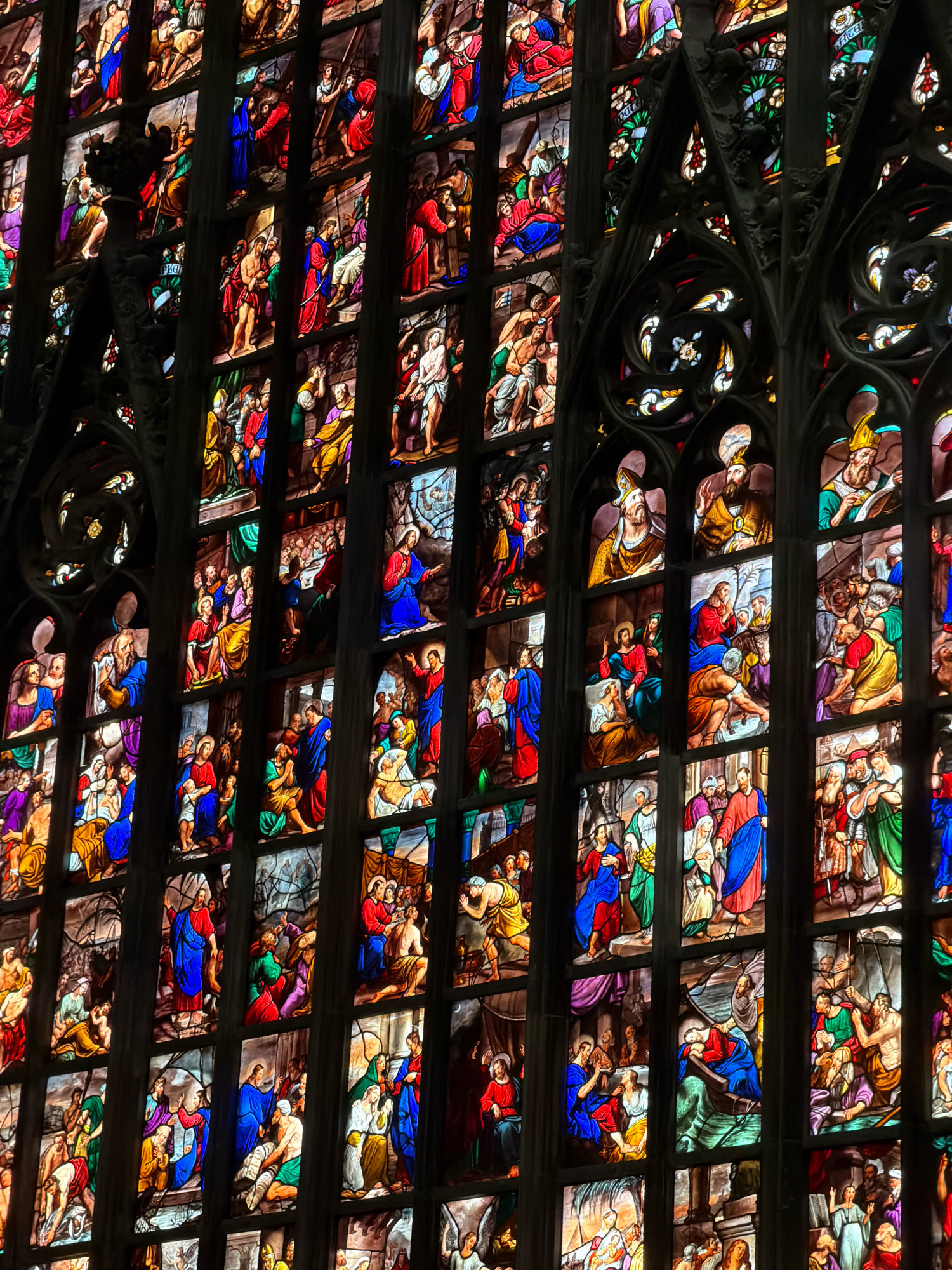 This is a detailed view of a stained-glass window within the Milan Cathedral (Duomo di Milano) in Milan, Italy. The window depicts various scenes from religious narratives, likely the life of Christ, rendered in vibrant colored glass. The artwork is divided into numerous rectangular panels, each showcasing multiple figures engaged in specific actions.
In the upper sections, there are depictions of figures in robes, some carrying or supporting others. One panel shows a figure on a donkey, accompanied by several people. Throughout the window, individuals are shown in poses of teaching, healing, or sorrow. Many figures are dressed in robes of red, blue, yellow, and white. There are scenes of people gathered around a central figure, some kneeling or looking upwards. In some panels, figures are shown being bound or led, suggesting depictions of suffering or persecution. The glass is set within intricate dark metal tracery that divides the panels and forms decorative elements, hinting at Gothic architectural style. The overall impression is one of narrative storytelling through religious art, with light filtering through the colored glass to illuminate the depicted events.