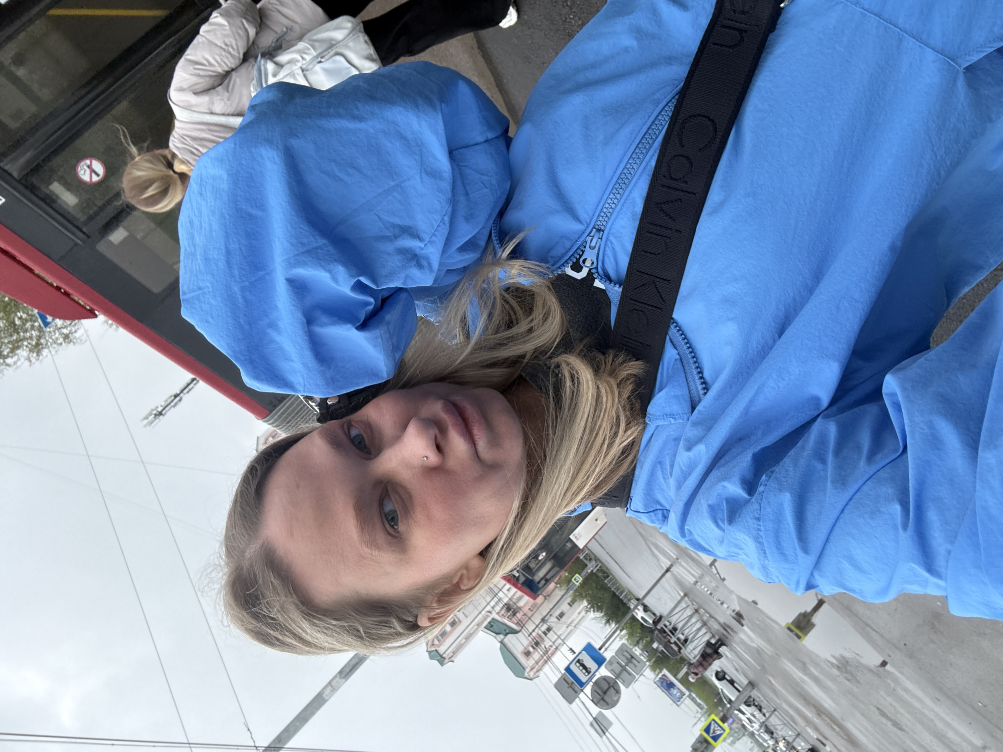 A young woman with blond hair and blue eyes stands on a sidewalk in the city of Perm, Russia. She wears a light blue hooded jacket with a black Calvin Klein strap across her body. A bus with a red accent is visible behind her. The weather appears overcast, and the street is wet, possibly from recent rain. A variety of street signs are visible in the background, along with buildings and cars. People are seen walking and riding in the bus.