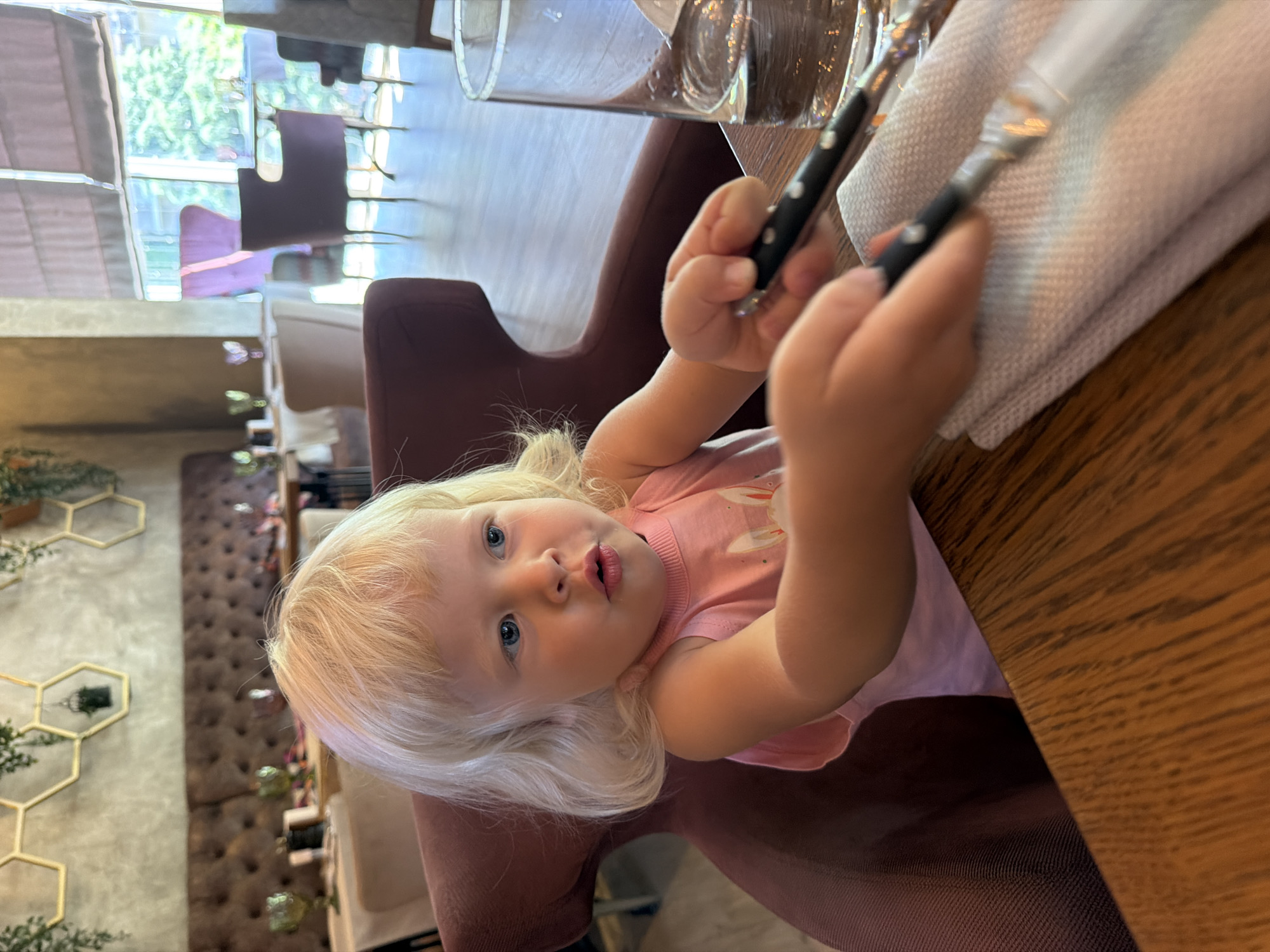 A young girl with blonde hair and blue eyes, wearing a pink tank top with a rabbit print, sits in a burgundy armchair in a restaurant. She is holding cutlery in her hands, appearing to interact with a napkin on a wooden table. In the background, the restaurant features plush seating, tables set with white tablecloths, and decorative hexagonal wall art. Large windows reveal a view of trees and other seating areas. The setting suggests a casual dining establishment, possibly in Sochi, Russia, given the context.