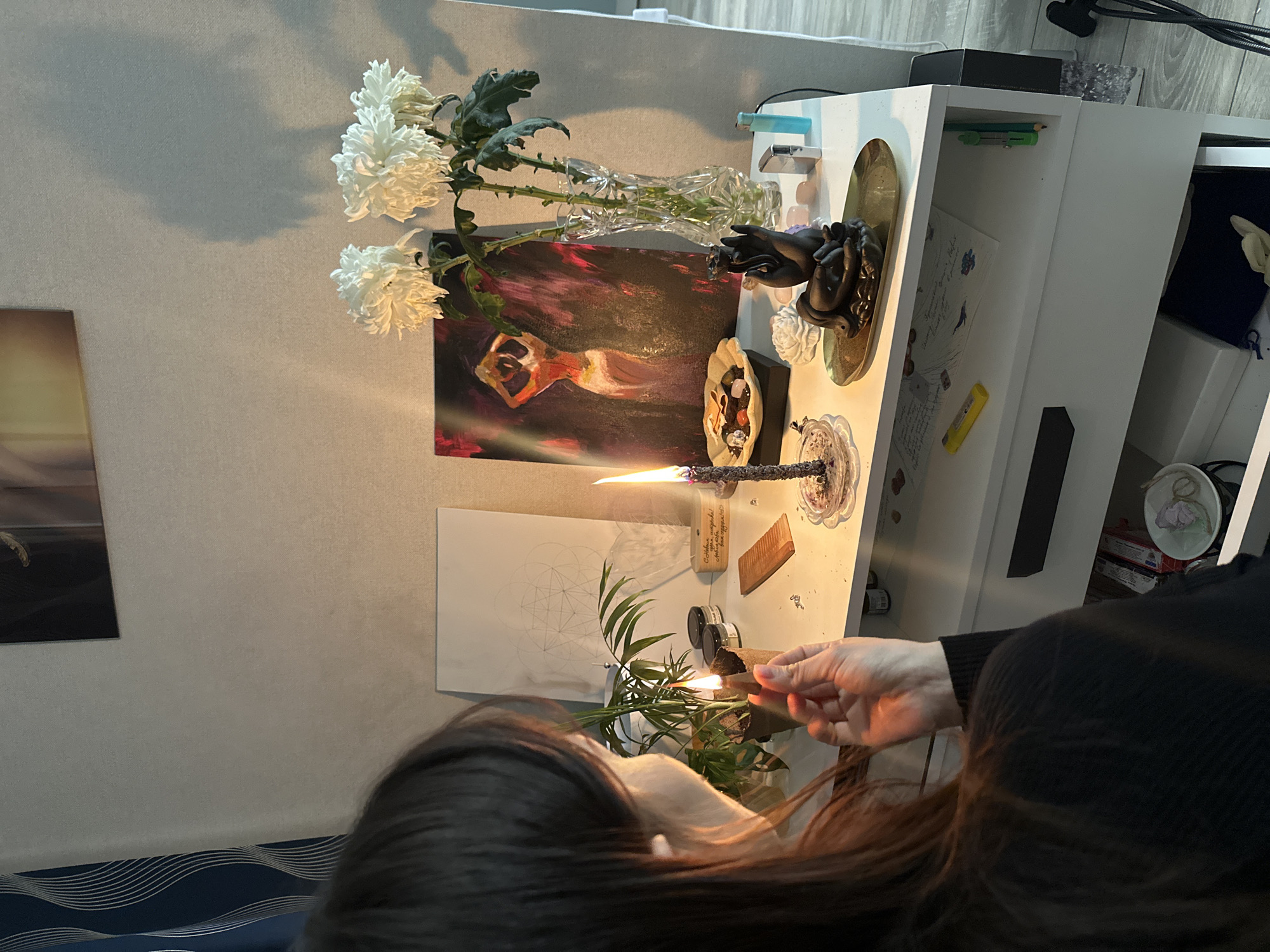 A woman with long brown hair is seen from above, sitting on a blue and white striped cushion. Her right hand is holding a lit rolled object, possibly incense, near a small potted plant with green leaves. The scene is situated on a white shelf unit that features various items, including:
* A painting with a figure depicted in motion.
* A vase holding white flowers with green leaves.
* A statue of a human figure resting its head on clasped hands.
* Various crystals and decorative stones.
* A lit incense stick in a holder.
* A piece of paper with geometric drawings.
* Two small jars.
The items are arranged on a shelf that appears to be located indoors, near a wall with a mounted painting. There is no indication as to whether the location is within the city of Murino, Russia.