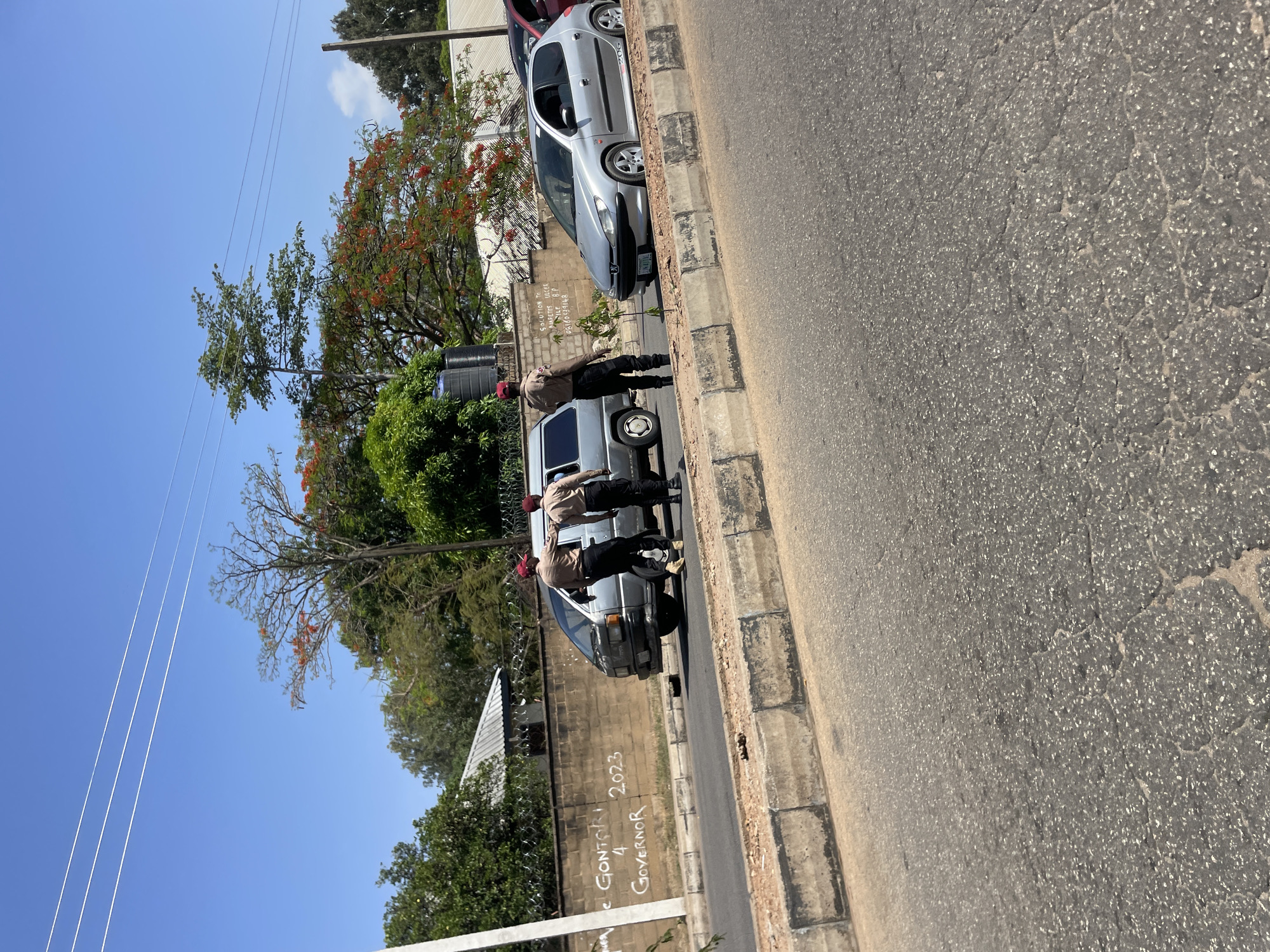 The image shows a street scene in Jos, Nigeria. Three men dressed in khaki uniforms and red caps stand alongside a parked silver minivan. They appear to be inspecting the vehicle. Behind them, a silver Peugeot 206 is also parked. A wall adjacent to the vehicles has graffiti that reads "El Gontoi 4 Governor 2023". The sky is clear blue. The road is paved and shows signs of wear. Trees and foliage are visible in the background.