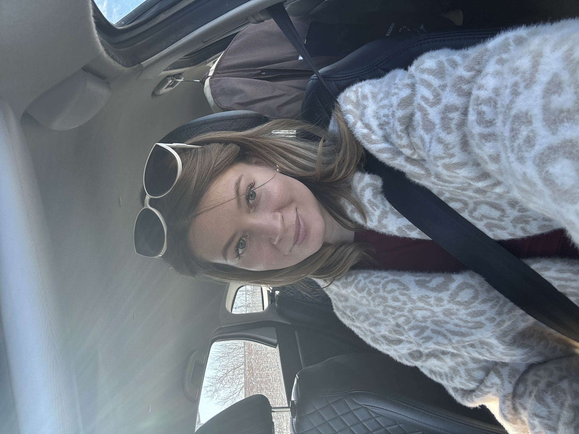 A woman wearing a seatbelt and a patterned sweater sits in the driver's seat of a car. Sunglasses rest on her head. Out the window, bare trees are visible, suggesting a late fall or winter environment. It is not possible to confirm the location as Chaikovsky, Russia from the given information.