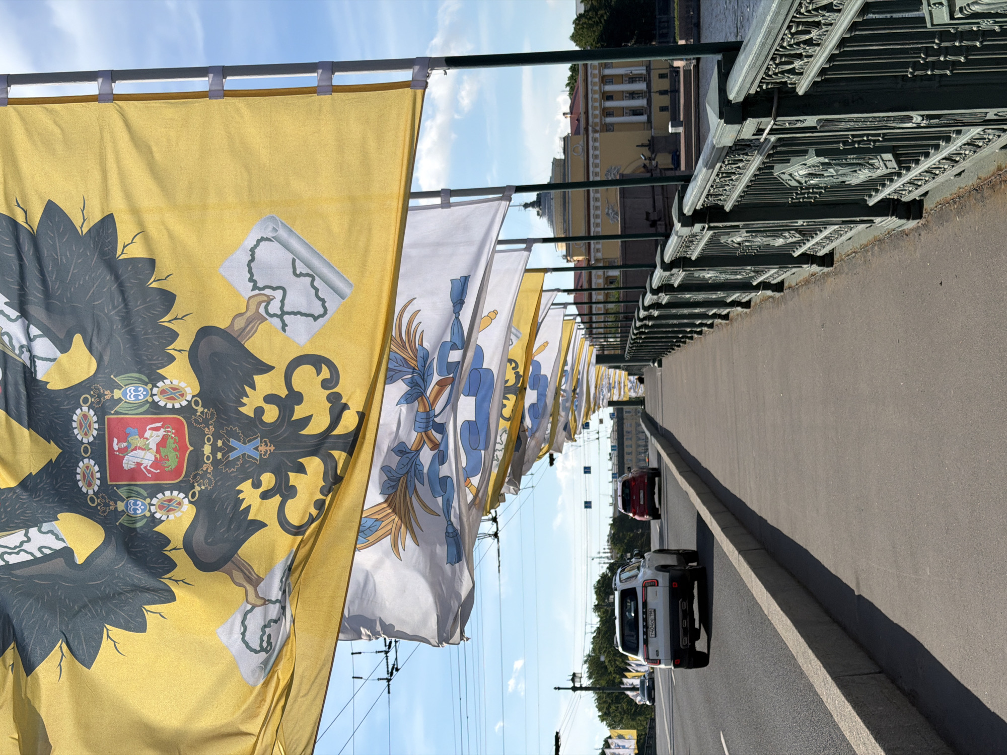 The image depicts a view from the Palace Bridge in Saint Petersburg, Russia, looking east. In the foreground, large yellow and white flags with elaborate designs, featuring double-headed eagles and coats of arms, are prominently displayed, attached to poles along the pedestrian walkway. The pedestrian walkway runs alongside a roadway with traffic. A white SUV and a red SUV are visible on the roadway, moving away from the viewer. In the distance, the Admiralty building, a landmark of Saint Petersburg, is visible with its distinctive spire. The sky is blue with scattered clouds. The overall scene conveys a sense of civic pride and architectural grandeur.